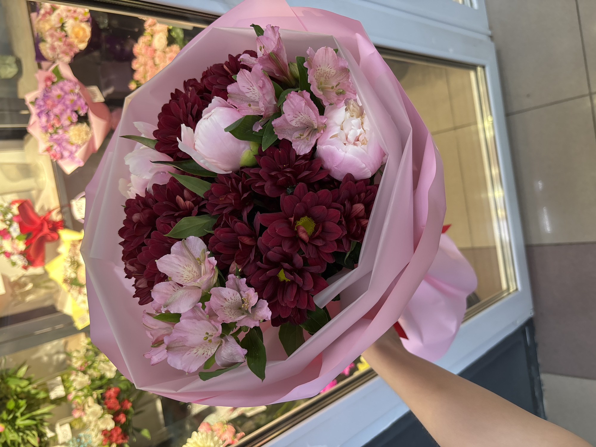 A bouquet of flowers, wrapped in pink paper, is held by a person's arm in front of a flower shop window. The bouquet contains burgundy chrysanthemums, pink peonies, and light pink alstroemerias. The shop window displays other flower arrangements, including pink and purple bouquets. The ground outside the shop is paved with beige tiles. The image appears to be taken in Chaikovsky, Russia, as indicated by the floral business sign.