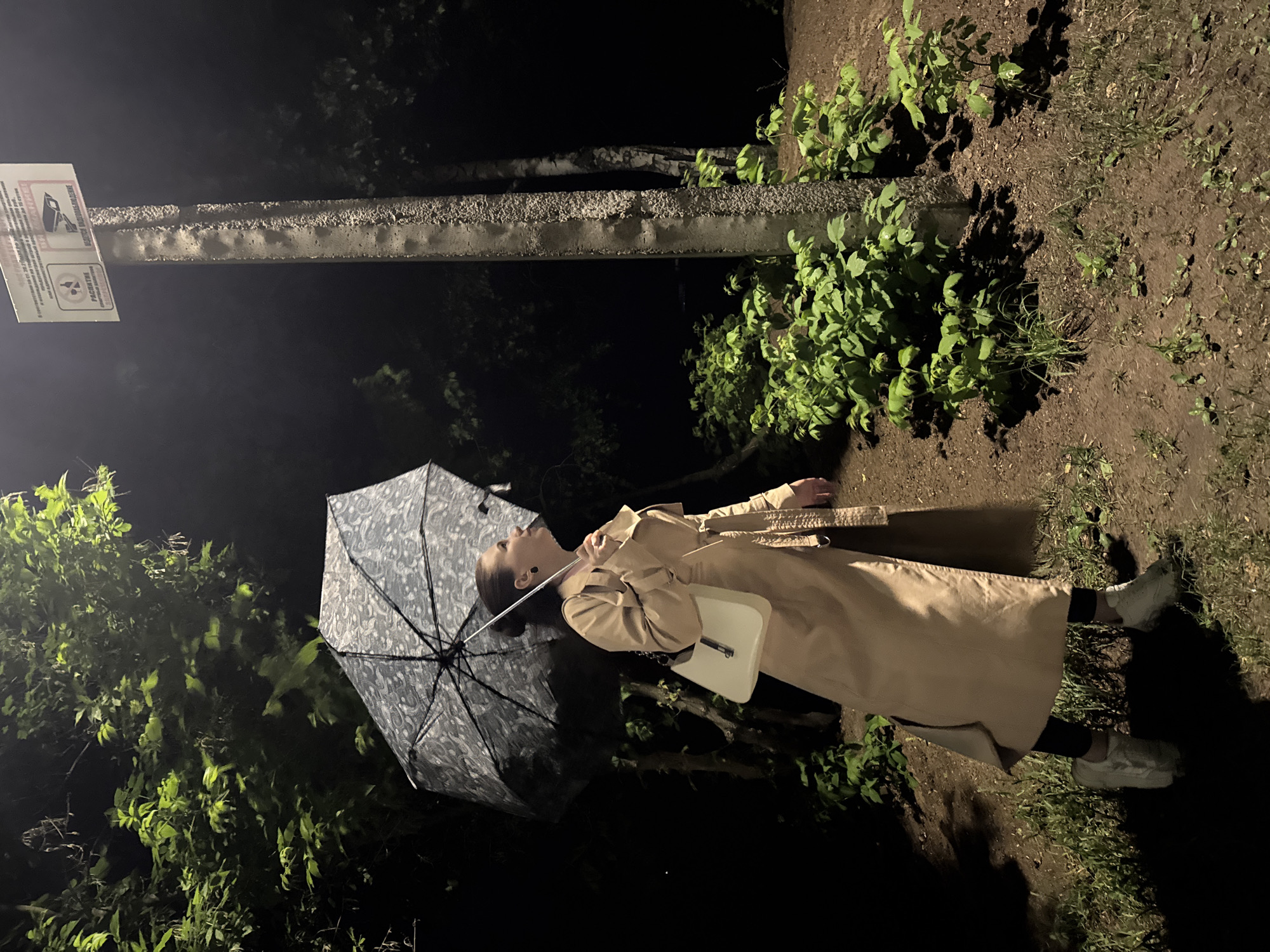 A female individual stands outdoors at night in Chaikovsky, Russia, illuminated by an artificial light source. She is wearing a long, light beige trench coat, black leggings, and white athletic shoes. Her dark hair is pulled back, and she has dark earrings. She holds an open gray umbrella with a light patterned design in her left hand and a white shoulder bag on her right side. Her gaze is directed upwards towards a textured concrete pole featuring two rectangular white signs. The right sign displays a surveillance camera icon and the Russian text "ВЕДЕТСЯ ВИДЕОНАБЛЮДЕНИЕ" (Video surveillance is being conducted). The left sign shows a crossed-out alcoholic beverage glass and the Russian text "РАСПИТИЕ СПИРТНЫХ НАПИТКОВ ЗАПРЕЩЕНО" (Consumption of alcoholic beverages is prohibited). The ground is composed of dark earth with patches of green grass and small, leafy bushes surrounding the base of the pole. In the background, dark foliage from trees, including birch trees on the right, is visible against the night sky. Strong shadows are cast on the ground due to the artificial lighting.