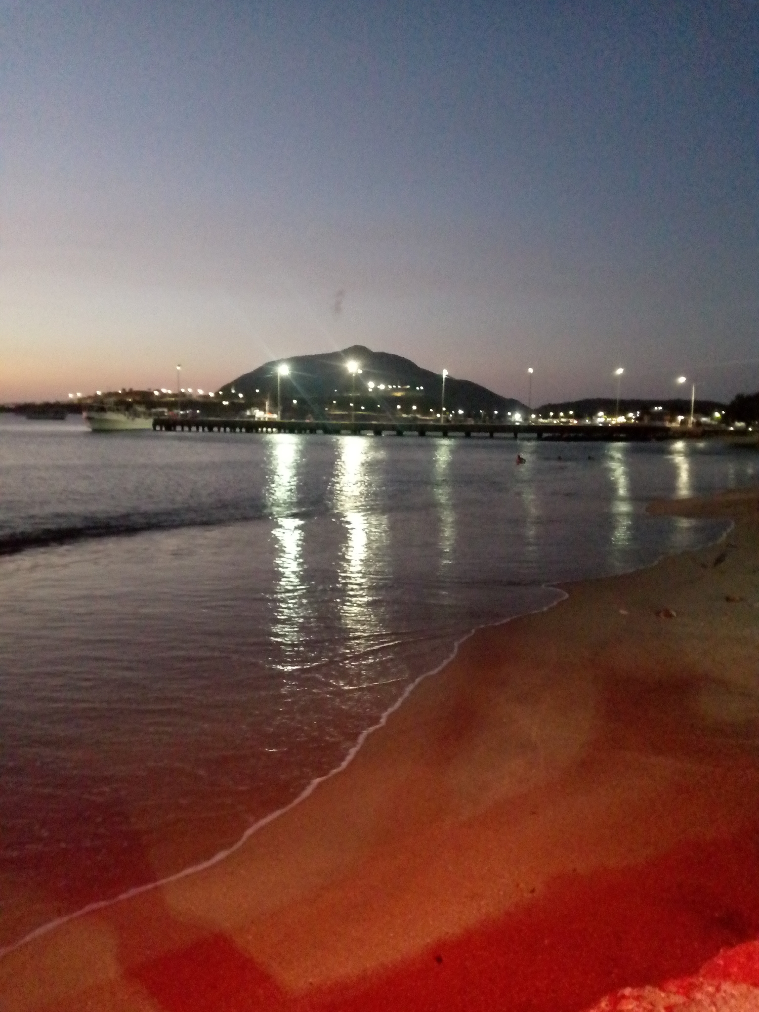 The image shows a tranquil beach scene in Juan Griego, Venezuela, at dusk. The foreground is dominated by the sandy beach and the gentle waves washing ashore. Beyond the beach, the water reflects the lights from a pier or walkway that extends into the bay. The pier is illuminated by a series of evenly spaced streetlights. In the background, a land mass, possibly a hill or small mountain, is silhouetted against the fading twilight sky. A single boat is docked near the pier.