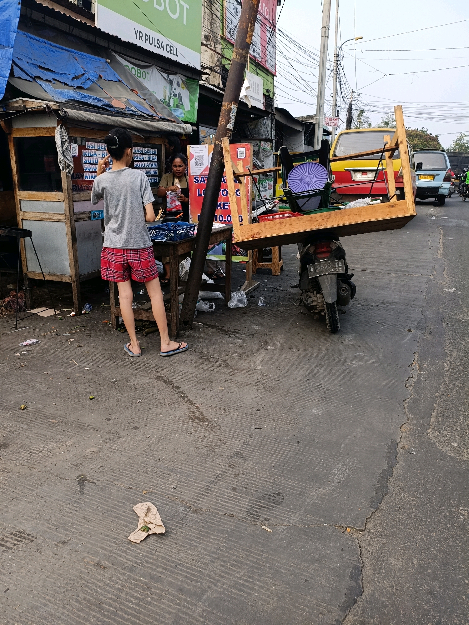 In Tangerang, Indonesia, on a paved outdoor surface, a person stands with their back to the camera, wearing a grey t-shirt and red plaid shorts, facing a food stall. The stall has blue tarpaulin roofing, wooden structures, and visible signs with Indonesian text. A woman in a dark top and apron is seen behind the counter, interacting with the person. In the foreground and to the right, a wooden structure, possibly a dismantled table or cart, is precariously balanced on the back of a parked motorcycle. The motorcycle's license plate is partially visible. The environment appears to be a street or roadside area, with other vehicles and buildings in the background, including a yellow and red van and a light blue van. Debris and trash are scattered on the ground.