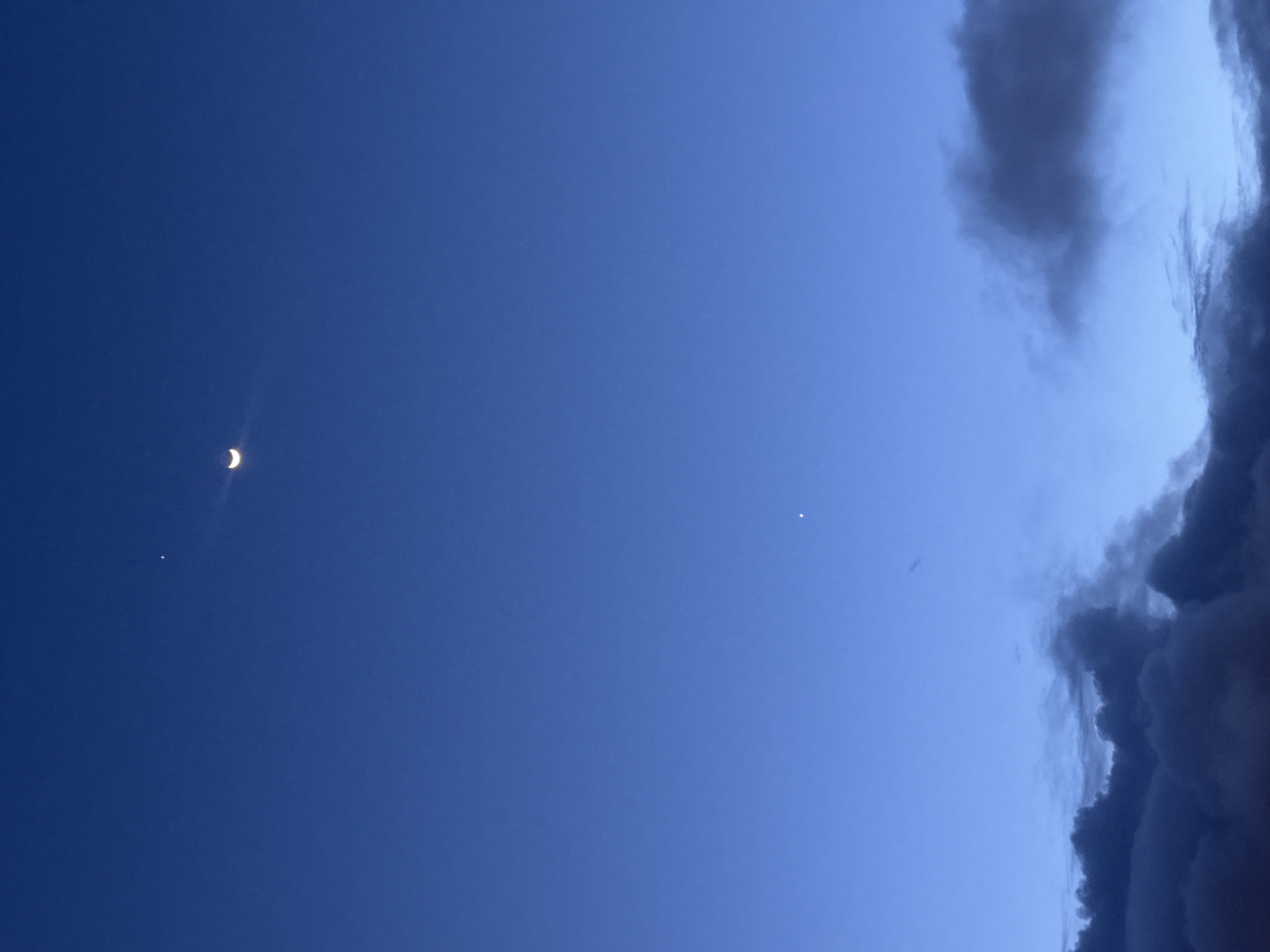 A twilight image shows a crescent moon in the upper left, two small stars below it, and a bank of dark clouds on the right side. The rest of the image is a dark blue sky. The image was taken at night in the city of Da Lat, Vietnam.