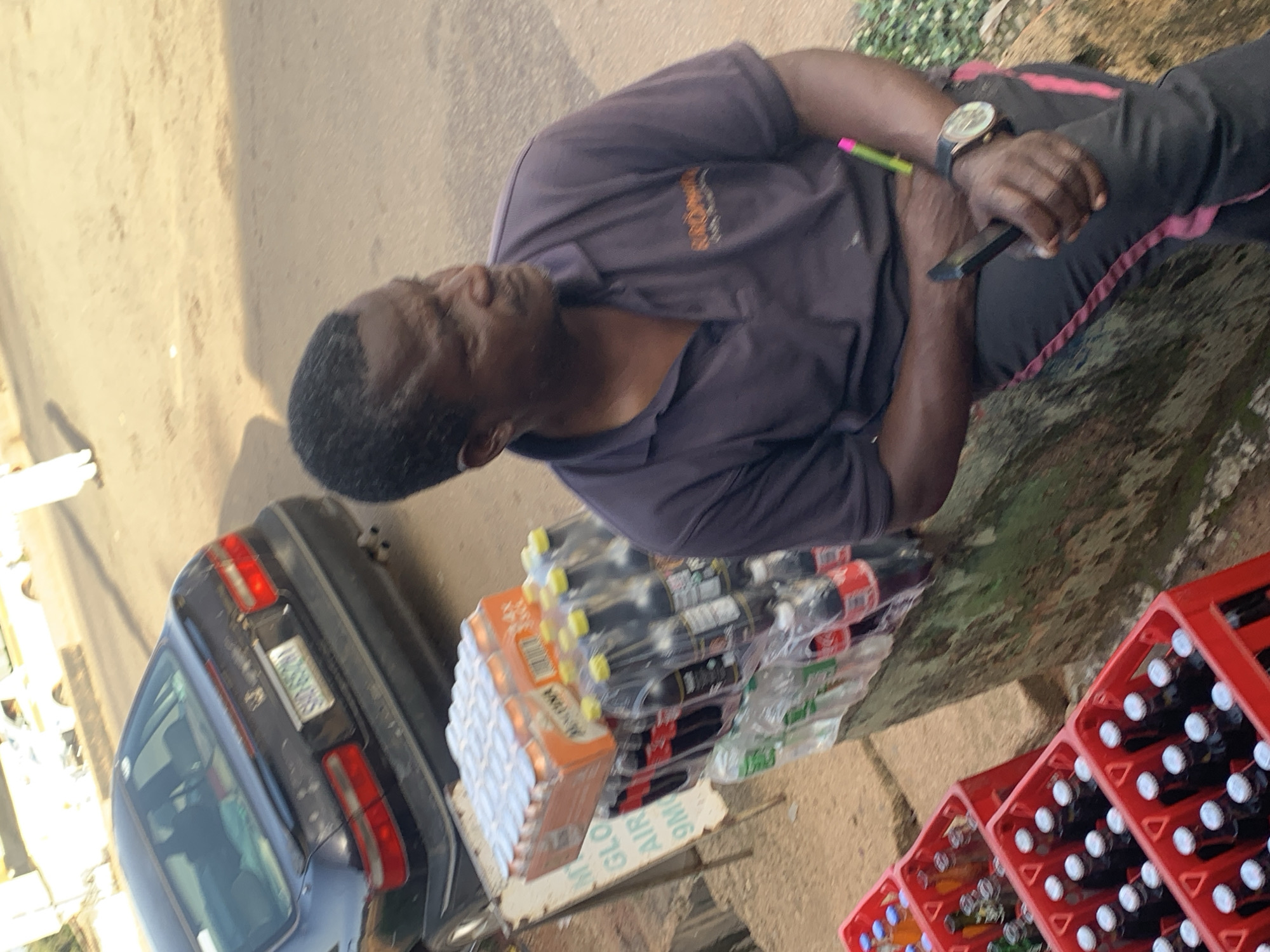 A man is standing outdoors, near a street, with a car parked behind him. He is wearing a dark purple polo shirt and dark pants with a pink stripe. He has a watch on his left wrist and is holding a pen in his right hand. Next to him, there are stacks of drinks, some in plastic bottles and others in red crates. The bottles are mostly dark colored, suggesting they might be sodas. There are also some orange boxes with white items inside. The scene appears to be a market or roadside stall. The ground is a mix of dirt and pavement. The lighting suggests it is daytime, possibly late morning or afternoon, and the weather seems to be clear. The man's expression is neutral, and he appears to be engaged in some activity with the pen and perhaps a notepad or register, which is partially visible. The overall setting suggests a commercial transaction taking place in a Nigerian urban environment. There is some visible text on the orange boxes, which appears to read "GLO AIRTIME 9Mobile".