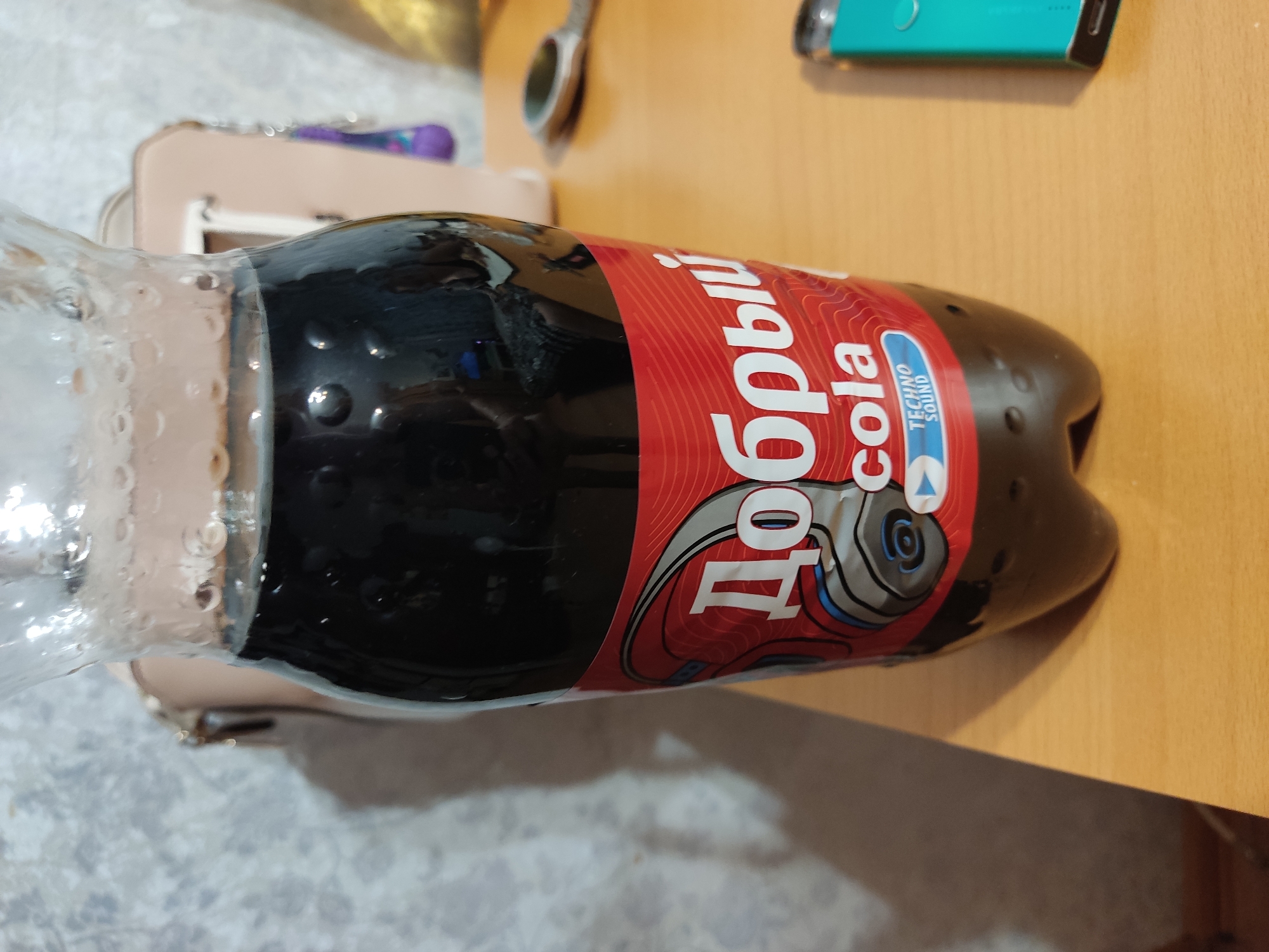 The image shows a close-up of a bottle of "Добрый Cola" with a red label featuring headphones and the text "TECHNO SOUND" on a wooden table. To the right, a teal-colored electronic device is visible. In the background, a light-colored purse and a grey, textured wall are present. There is no indication of the location being Serpukhov, Russia, or any other specific city. The scene is static and does not depict any people or actions.