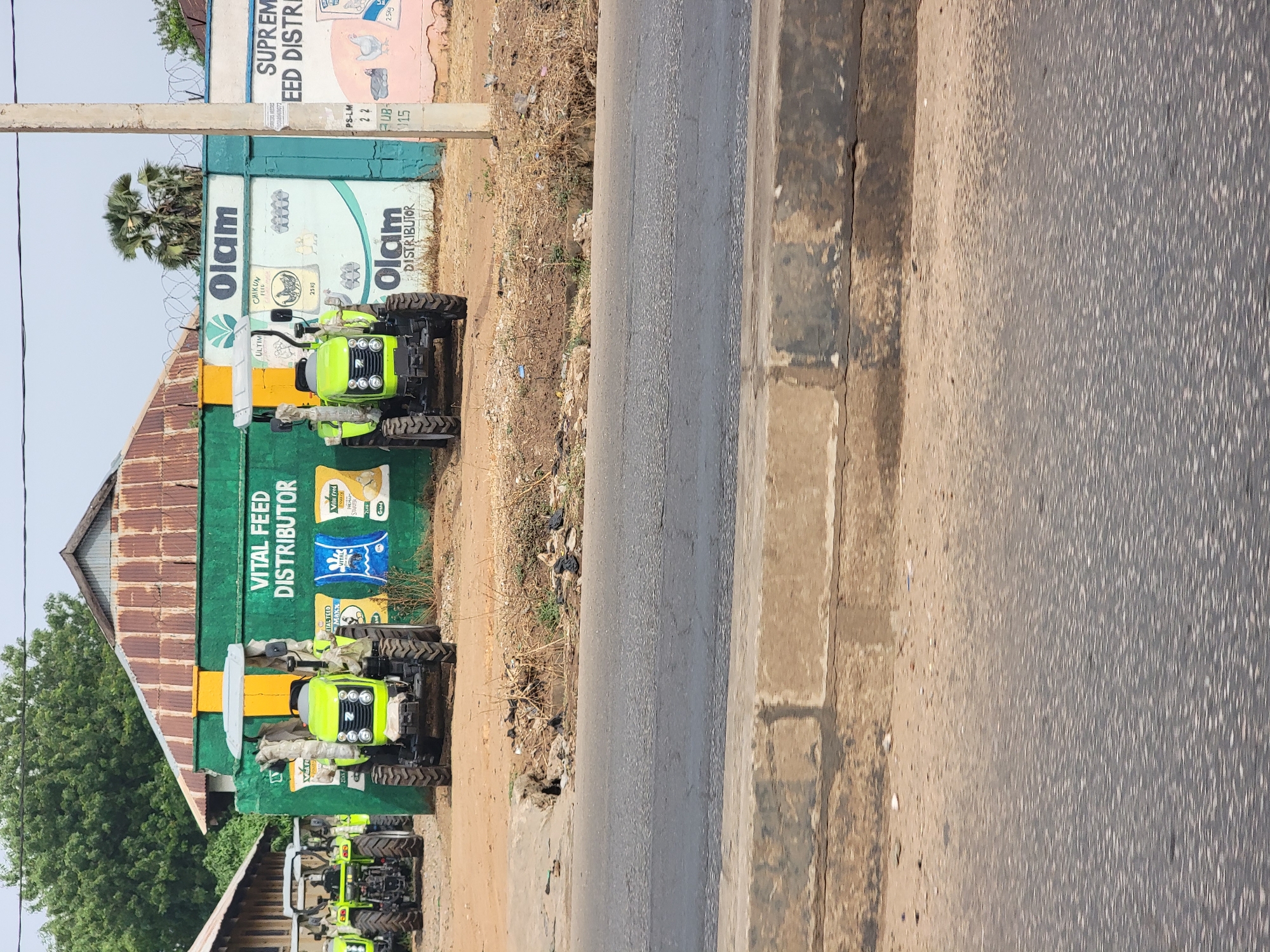 The image depicts a row of green tractors parked in front of a building with advertisements in what appears to be Barnawa, Nigeria. The building is painted with "VITAL FEED DISTRIBUTOR" in white letters and features images of animal feed products. The name "Olam" is prominently displayed on the building as well. The tractors are new and appear to be for sale or distribution. A paved road runs along the edge of the scene. Additional signage for "SUPREME FEED DISTRI" is partially visible. There are no people visible in the immediate scene. The environment appears to be a commercial area.