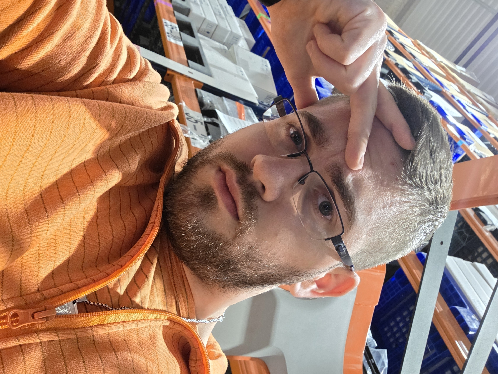 A close-up selfie shows a man with light brown hair and a beard/stubble, wearing an orange hooded sweatshirt or jacket with a partially unzipped front and a visible silver chain around his neck. He is wearing glasses, and his right hand is raised, with fingers touching his forehead above his right eye, possibly as a gesture or for support while taking the photo. His expression is neutral to slightly contemplative.
The background reveals an indoor setting, likely a warehouse or storage facility, characterized by industrial shelving and racks. These shelves are filled with various items, including multiple white boxes and blue plastic crates or containers. Some shelves appear to be orange. On one of the orange shelf labels, the numbers "65-30" are visible. The lighting is bright, suggesting either strong artificial lights or overhead natural light. The zipper on the man's jacket has a small, indiscernible emblem on the pull.