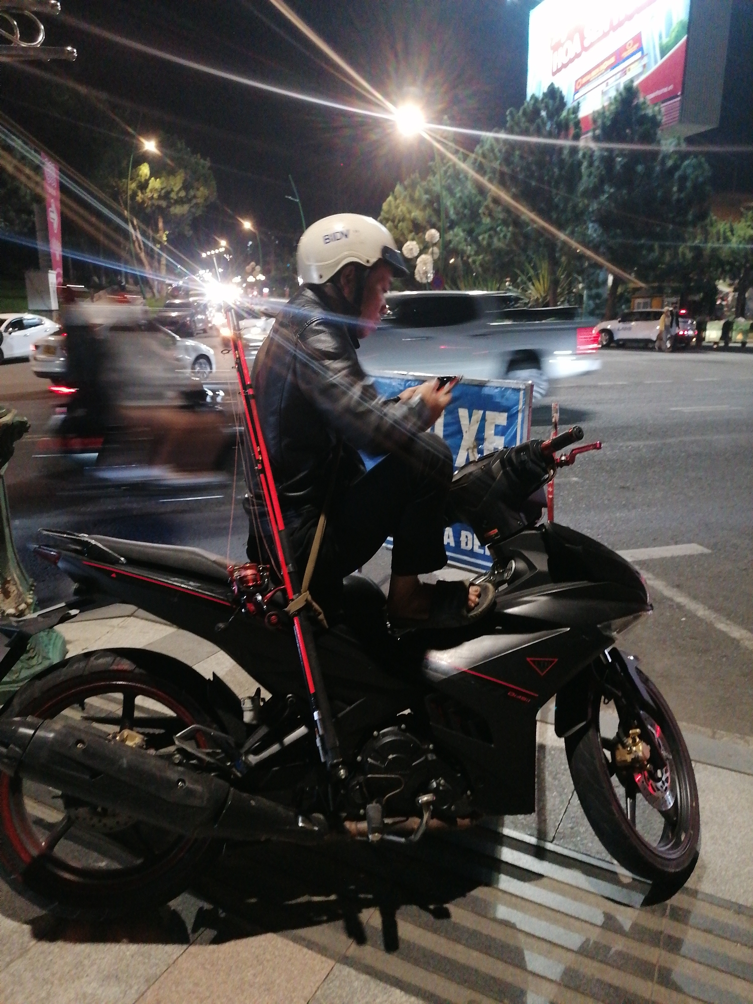 A person wearing a white helmet is riding a black motorcycle at night in Da Lat, Vietnam. The rider is looking at their phone and has two fishing rods strapped to the side of the motorcycle. The motorcycle is black with red accents and has a large exhaust pipe. The background shows a street scene with other vehicles, streetlights, and trees. The rider is wearing a dark jacket and dark pants. The environment appears to be an urban area.