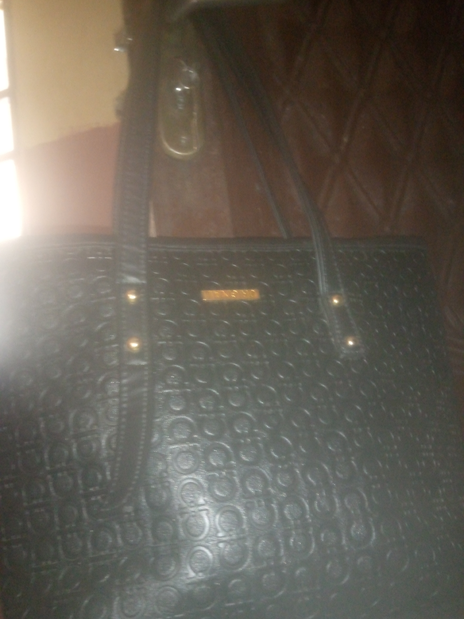 A black tote bag with a patterned exterior is displayed against a dark, textured background. The bag has two shoulder straps, one of which is visible on the left side of the image, and two gold-toned metal studs are present on the front of the bag. A gold-toned logo plate with text is centered on the front of the bag. The background appears to be a wooden surface, possibly a wall or furniture. The lighting in the image is uneven, with a bright light source to the left casting glare and shadows. The location in the city of Unknown, Unknown is not determinable from the image.