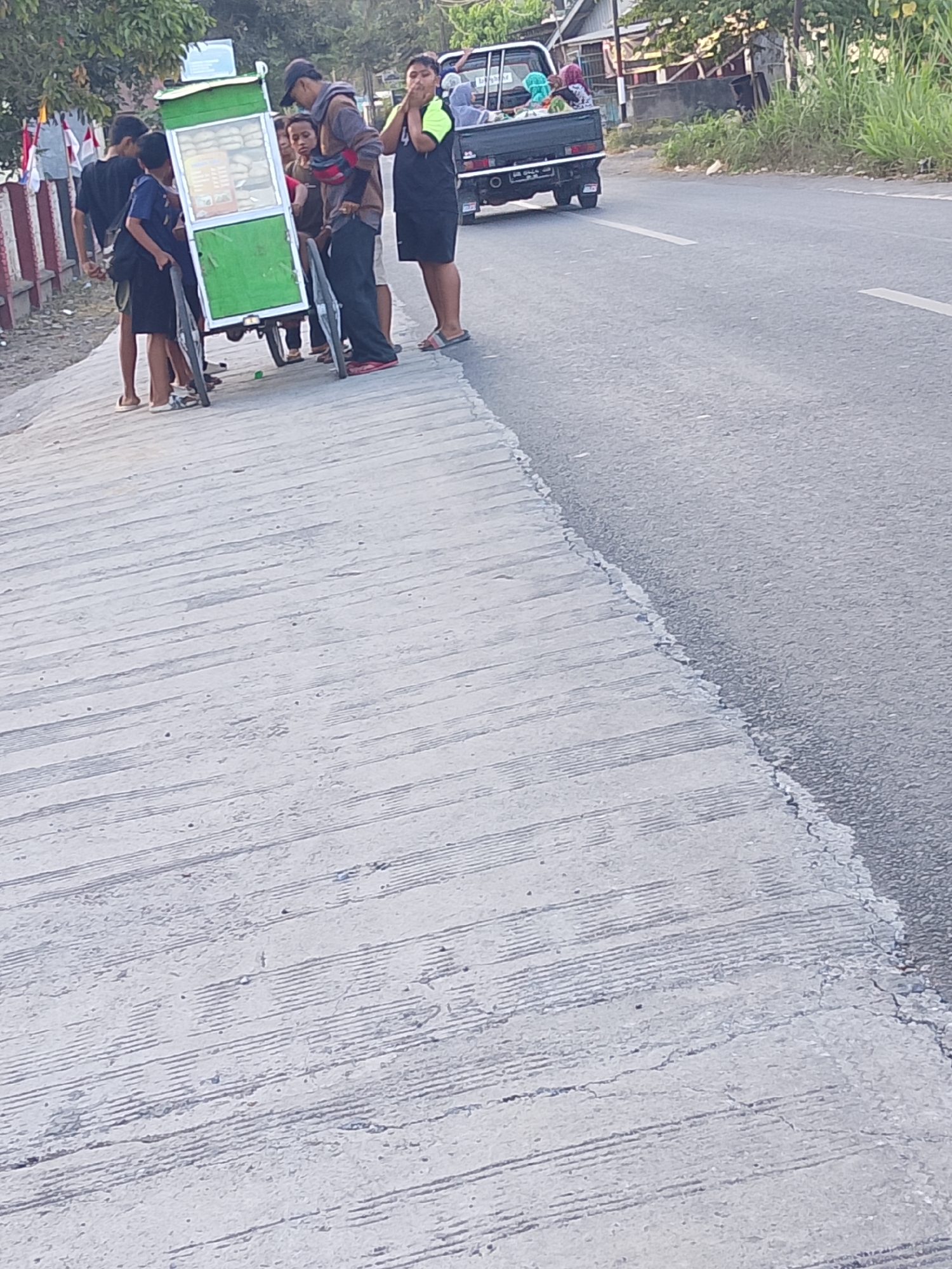 In an outdoor scene with a cracked concrete surface on the left and asphalt on the right, a group of people stands around a green and white food cart. The cart has wheels and appears to be filled with goods. Several individuals, including young males and at least one adult male wearing a cap, are gathered near the cart. One person, possibly a young male, is looking towards the cart with his hand near his face, while another, a young male in a black shirt and shorts, stands to the left of the cart. An adult male in a patterned shirt and dark pants stands behind the cart, holding something red and black across his chest. To the right of the cart, another young male wears a black and lime green shirt and dark shorts. In the background, a dark-colored pickup truck is visible on the asphalt road, with other people in its bed. The environment suggests a roadside setting, possibly in a rural or suburban area, with some vegetation and buildings in the distance.