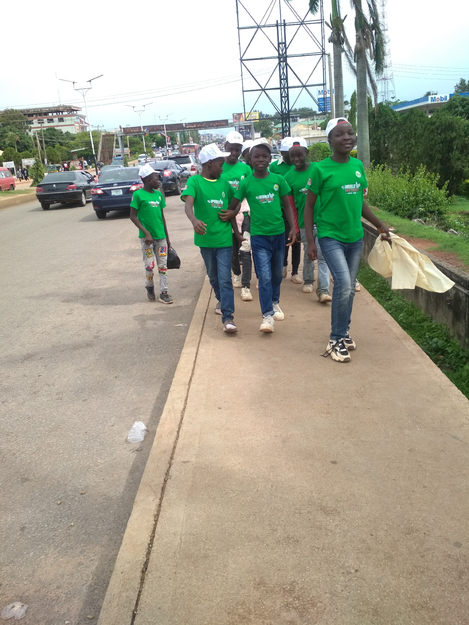 This image depicts a group of young people, likely children or teenagers, walking along a concrete sidewalk next to an asphalt road in an urban or suburban area, consistent with Jos, Nigeria. At least eight individuals are visible, all dressed in matching bright green t-shirts with a logo, blue jeans, and white baseball caps. Their uniform attire suggests they are participating in an organized event, such as a community march or public awareness campaign. The person in the foreground on the right is holding a light-colored piece of fabric, possibly part of a banner or flag.
The road beside the sidewalk has moderate traffic, with several cars including a black Peugeot and a blue sedan. In the background, the scene reveals typical urban infrastructure: various buildings, overhead power lines, tall streetlights, a large metal lattice structure (possibly a transmission tower or billboard), and a pedestrian bridge in the distance. To the right, a "Mobil" gas station sign is clearly visible, indicating a commercial zone. Palm trees, lush bushes, and green grass line the roadside.
The time of day appears to be daytime, under an overcast sky, resulting in bright yet diffused lighting without strong shadows. The expressions of the young people in the foreground range from neutral to smiling, suggesting a positive or engaged mood. On their green t-shirts, the text "WORLD CHILDHOOD" is partially legible, indicating the focus or theme of their activity. A plastic bottle or bag can be seen as litter on the road adjacent to the sidewalk.
