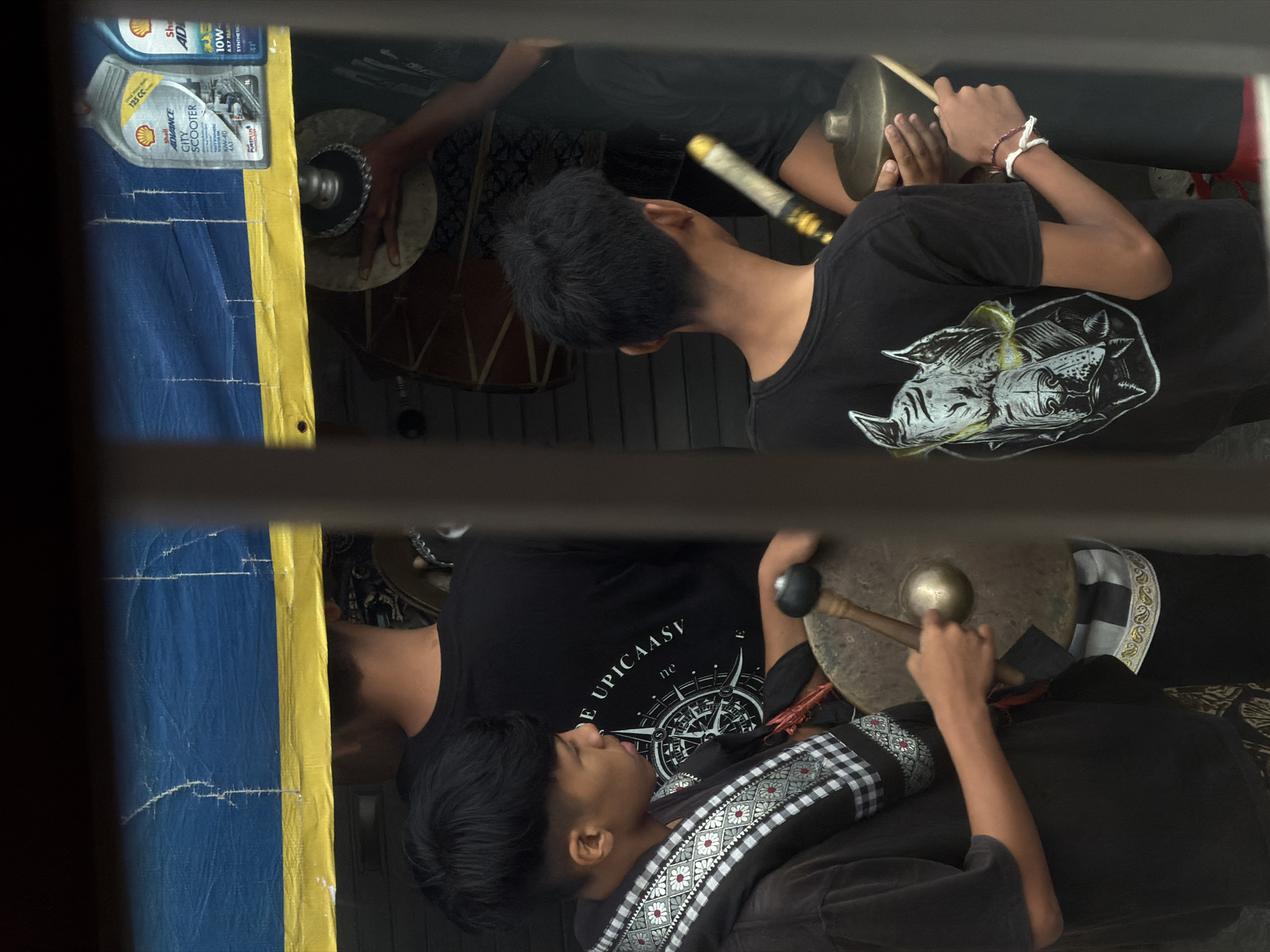 The image depicts a close-up, slightly blurred view of two young males in the process of playing traditional musical instruments, likely gongs, as part of a performance or cultural event. The setting appears to be outdoors, with a blue and yellow tarp providing shelter overhead. On the tarp, two bottles of Shell Advance scooter oil are visible, suggesting proximity to a commercial area or possibly sponsorship.
The male on the left is wearing a dark t-shirt with a black and white checkered and patterned scarf draped over his shoulder. He is holding a mallet in his right hand, poised to strike a large, bronze gong that is partially visible. His left hand rests near the gong. He has short, dark, styled hair.
The male on the right is wearing a black t-shirt with a graphic of a stylized dog's head. He is holding two drumsticks, one in each hand, and is also positioned to play instruments that are not fully visible. He has short, dark hair. His left wrist has two thin bracelets.
The overall scene is framed by what appears to be a window or a gap in a structure, adding a sense of looking into the scene. The lighting is subdued, contributing to the slightly muted colors and textures. The environment suggests a street-side setup or a temporary stall in North Kuta, Indonesia, where such performances are common during local festivals or gatherings. The presence of the oil bottles might indicate a location near a street or a business that also serves as a venue for cultural activities.
