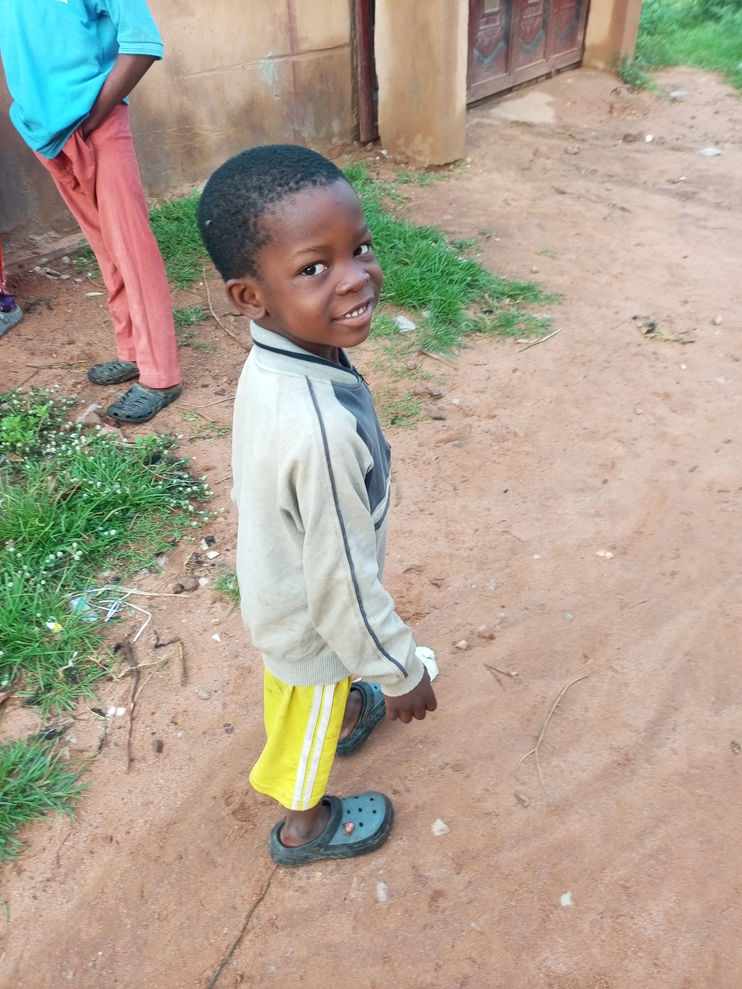 A young Black boy, smiling, stands on a dirt path in Tempure, Nigeria, looking over his right shoulder. He wears a light-colored jacket with dark accents and bright yellow shorts with white stripes. He is also wearing dark clogs. Behind him, a person in a blue shirt and rust-colored pants stands with their hands in their pockets. The ground is a mix of dirt and sparse green grass. In the background, there is a building with a large wooden door and a textured wall. The overall impression is of a candid outdoor shot in a rural or semi-urban setting.