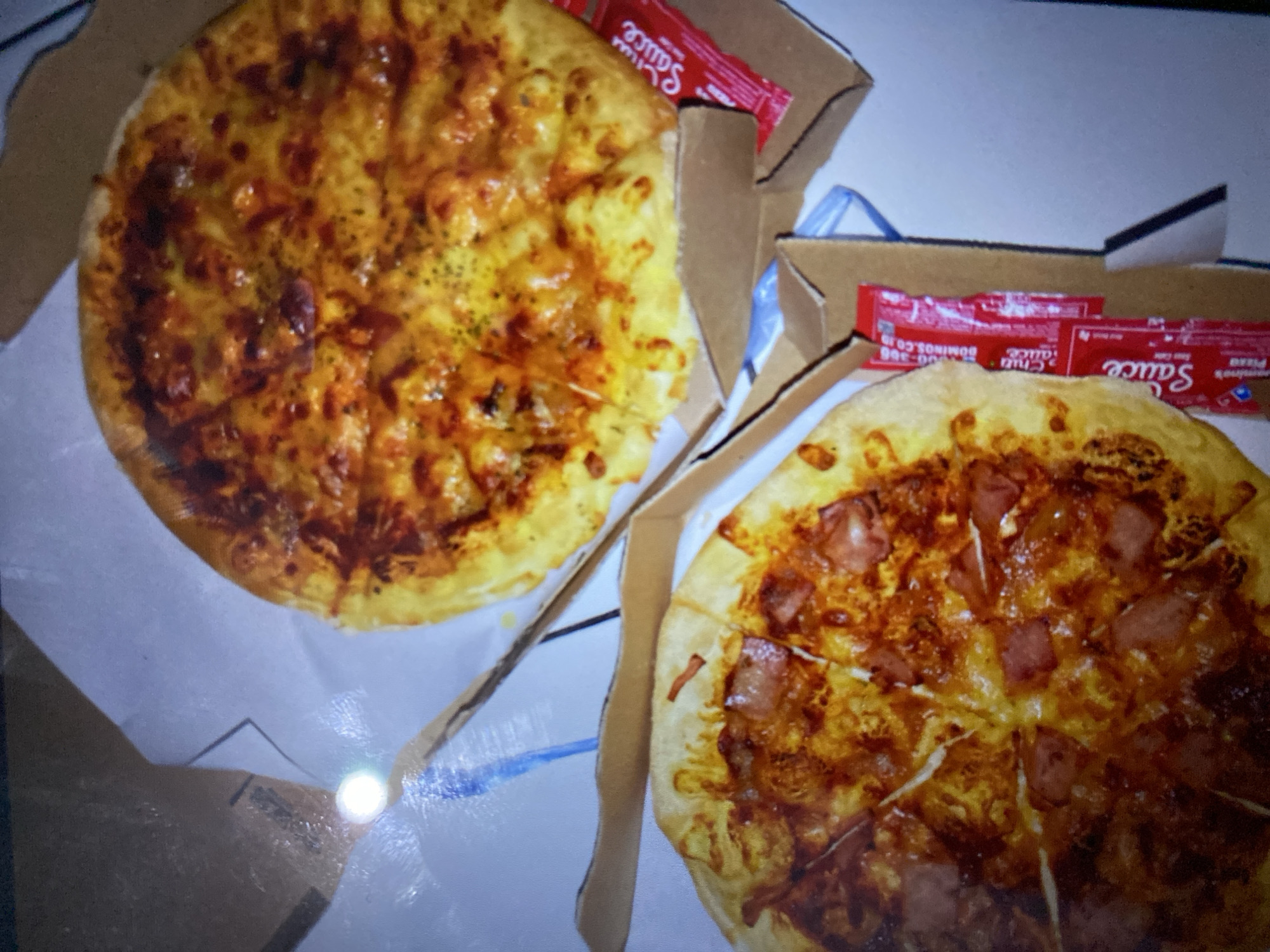 Two pizzas are presented in open cardboard pizza boxes on a white surface, possibly a table or counter. One pizza, located in the upper right, is topped with melted cheese and appears to have some herbs sprinkled on it. The other pizza, in the lower left, is topped with cheese and circular slices of meat, likely ham or pepperoni. Both pizzas are cut into slices. Adjacent to the pizzas are packets of red sauce, some labeled "Chili Sauce" and others with "Sauce" and "Domino's Pizza." A bright, circular reflection, likely from a light source, is visible on the white surface in the upper left quadrant of the image, near a folded brown cardboard edge. The lighting in the image is somewhat dim, and the overall scene suggests a casual meal setting. The location in Kalitinggar, Indonesia, is not visually evident from the provided image.