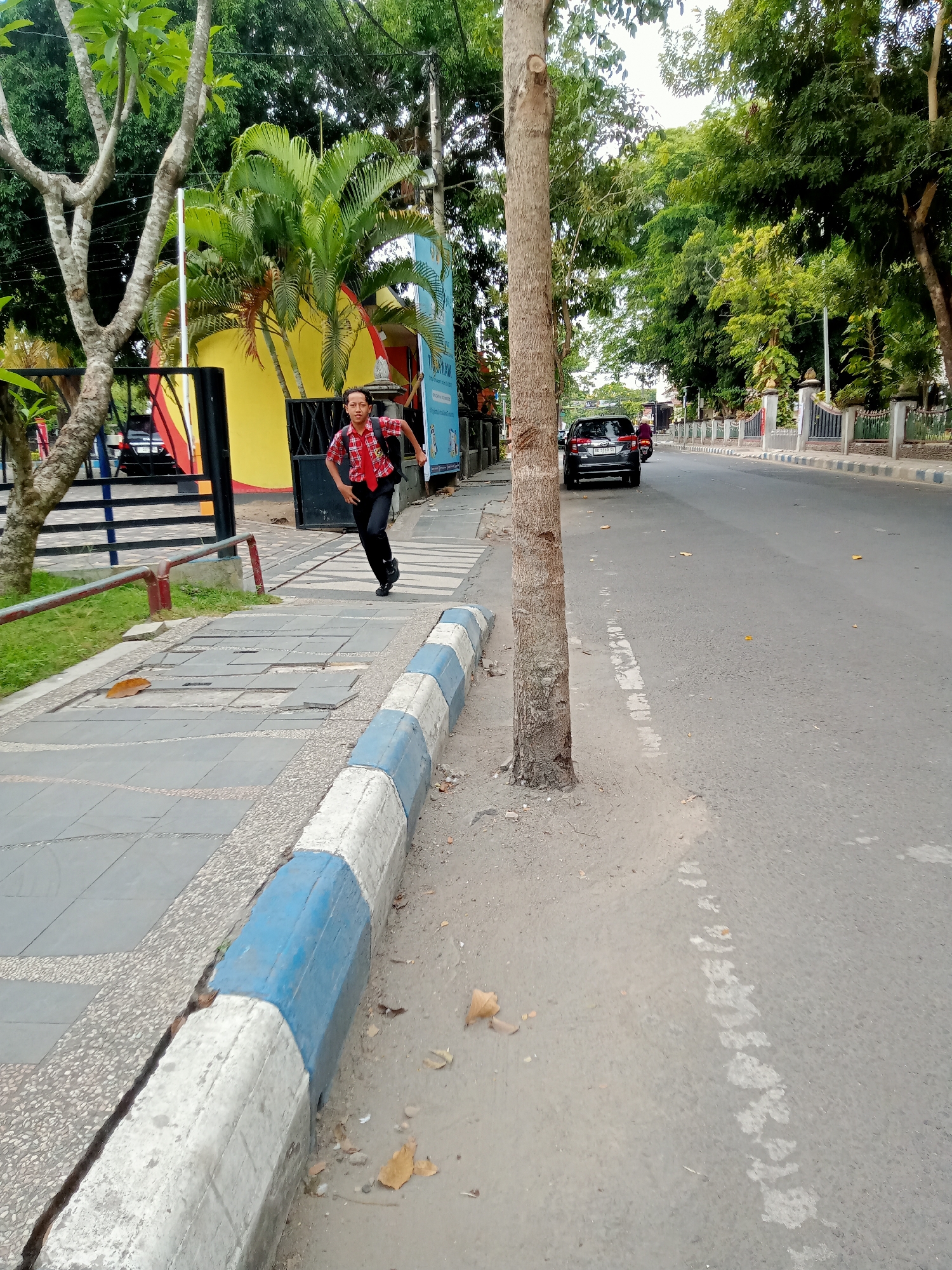 A young male wearing a red and black plaid shirt, black pants, and a backpack is captured in motion on a sidewalk next to a tree. The sidewalk is paved with grey tiles and bordered by a concrete curb painted with alternating blue and white sections. In the background, a yellow building with a black fence is visible, along with palm trees and lush green foliage. A street runs parallel to the sidewalk, with a black car and a motorcycle on it. The scene appears to be on a street in Tulungagung, Indonesia, possibly near a school or public institution given the presence of the colorful building and manicured trees.