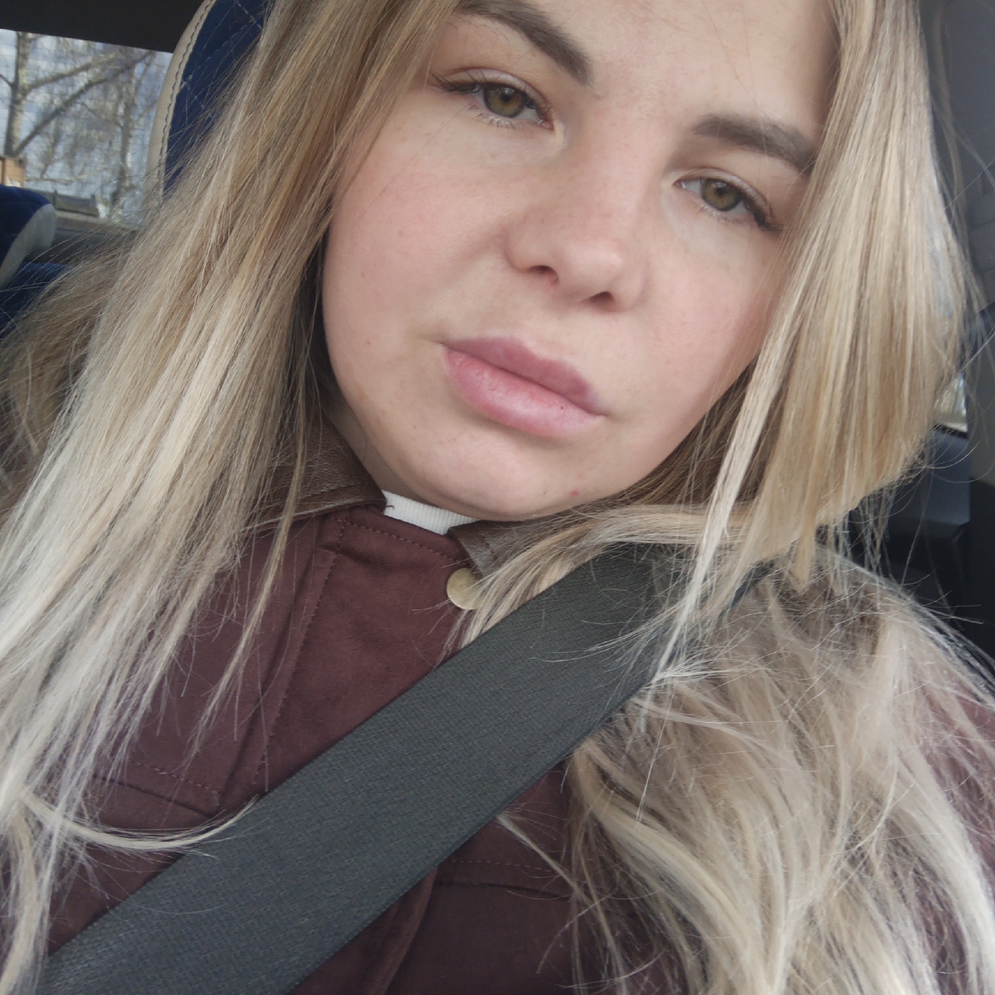 A blonde woman is in a car, wearing a maroon suede jacket and a grey seatbelt. The location is unknown. Trees are visible outside the car window.