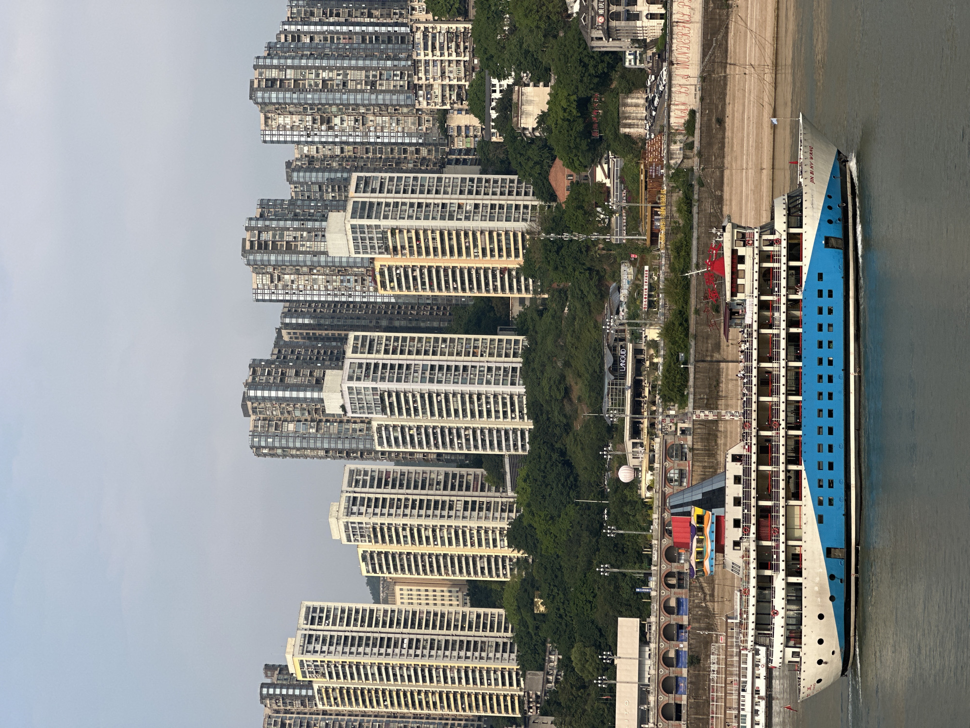 The image depicts a scene in Yuzhong District, China. On the left are dense clusters of high-rise apartment buildings. The buildings vary in architectural style but generally consist of stacked, rectangular units. A strip of green foliage divides the buildings from a concrete pathway running along a riverbank. A large, multi-deck river cruise ship is docked along the pathway, with its blue and white exterior facing the camera. The river occupies the right side of the frame.