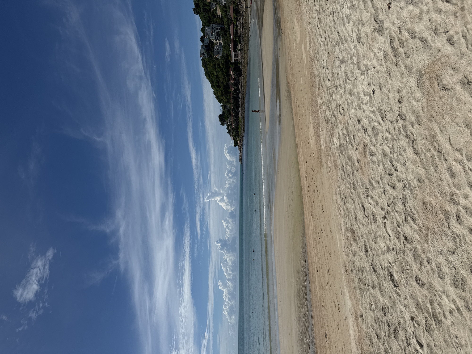 This is a photograph of a beach scene in Baan Chaweng Noi, Thailand. The image is oriented vertically. The foreground is dominated by a sandy beach, showing footprints and ripples from the receding tide. To the left of the beach, the calm, shallow ocean is visible, with a few small waves breaking gently. Further left, the sea transitions to a deeper blue under a bright sky filled with wispy white clouds and some more defined cumulus clouds.
Along the left edge of the scene, a steep, green, treed hillside rises, dotted with white buildings, suggesting houses or villas built on the incline. There are a few people visible in the water, some appearing to be swimming or wading, and at least one person walking along the shoreline. The lighting suggests it is daytime, likely late morning or early afternoon, with a clear and sunny sky. The overall mood of the image is peaceful and relaxing, capturing a typical tropical beach day. There is no visible text in the image.