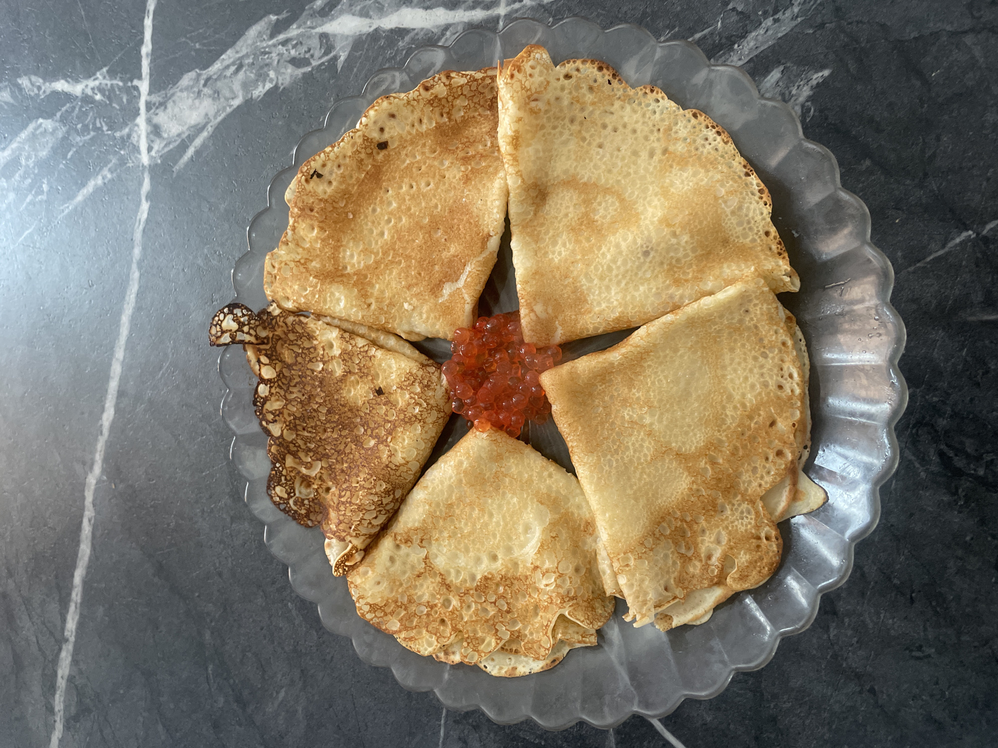 The image depicts a clear glass plate with scalloped edges, placed on a grey marble surface. Arranged on the plate are five folded crepes, positioned in a circle. At the center of the circle, there is a small pile of orange-red caviar. The crepes vary in color, with one appearing darker and more browned than the others. The setting appears to be indoors, potentially in a home or restaurant environment. No people are visible in the image. There is no information to determine if the image was taken in Baranovka, Russia.