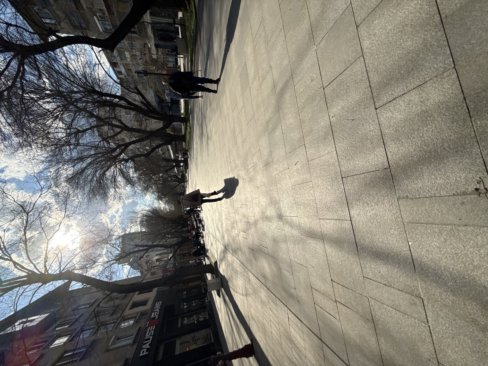 The image shows a pedestrian zone in Stara Zagora, Bulgaria, characterized by a paved walkway made of large, light-colored stone slabs. There are several pedestrians. One person with a long shadow is walking in the sun, and another person is walking further away. The sun is bright and low, casting long shadows from the people and trees. Bare trees line the street, and buildings with visible storefronts are adjacent to the walkway, with a sign for "PAUSE JEANS" visible. There are metal bicycle racks next to the trees and stores.
