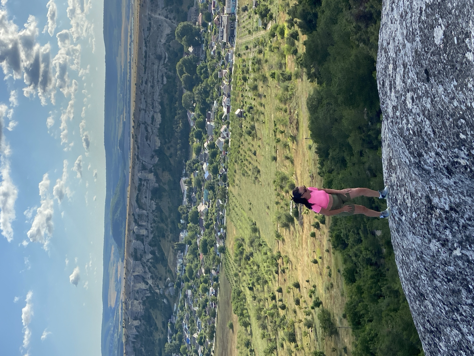 A woman stands on a large, textured rock in the foreground, looking downwards. She is wearing a bright pink t-shirt, khaki shorts, and athletic shoes. Her dark hair is pulled back. Behind her, a valley unfolds with a village nestled amongst green fields and trees. In the distance, a large, layered rock formation stretches across the horizon, with rolling hills beyond. The sky is blue with scattered white clouds. This scene is located in Krasnomakskoye rural settlement, Ukraine.