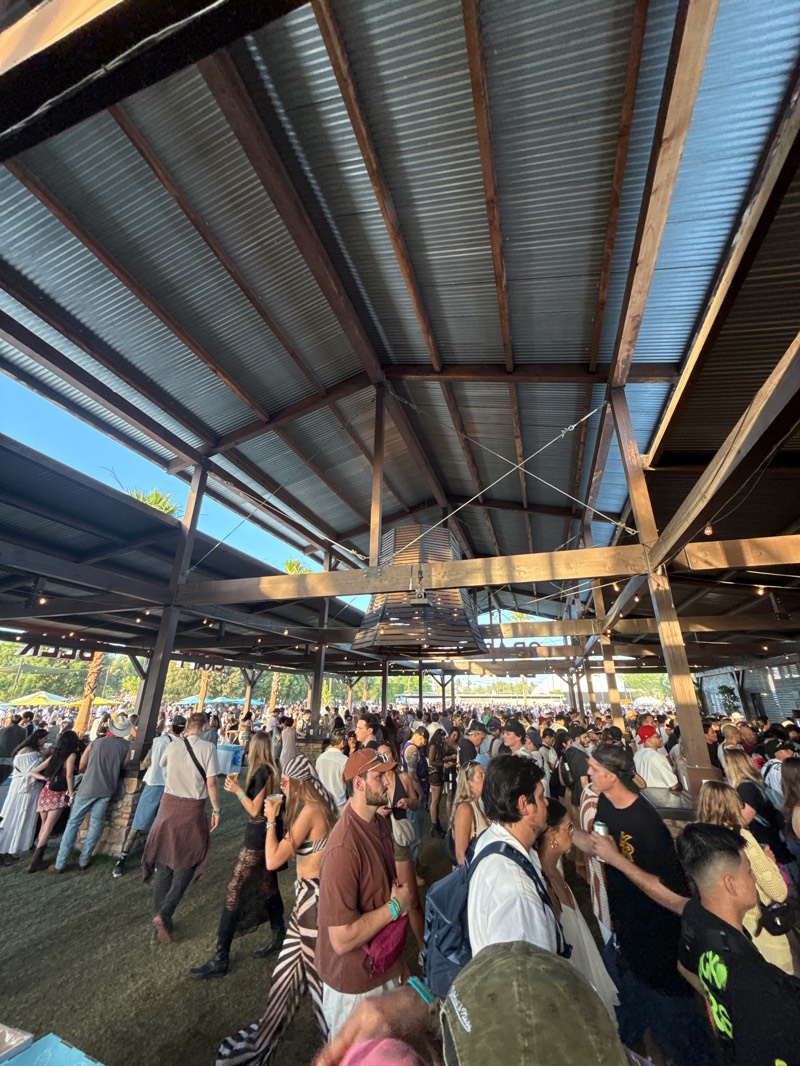 The image shows a large crowd of people gathered in an open-air venue with a corrugated metal roof supported by wooden beams. The location is Indio, United States, based on the likely setting of the Coachella music festival. People are dressed in a variety of casual clothing, and the overall atmosphere appears lively and crowded.