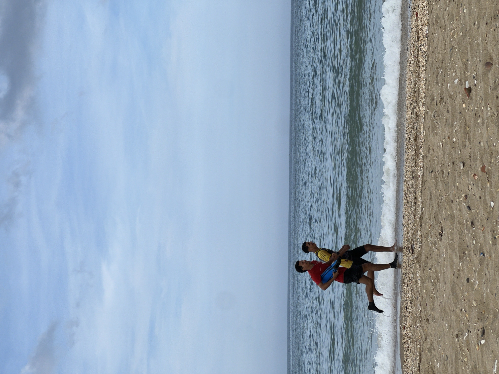 Two individuals are standing at the edge of the water on a beach. One is wearing a red shirt and carrying a blue object, the other is wearing a yellow shirt. The location is M'Diq, Morocco, as evidenced by the beach setting and coastal characteristics. The sky is overcast. Waves are visible at the shoreline. The beach consists of sand and small shells.