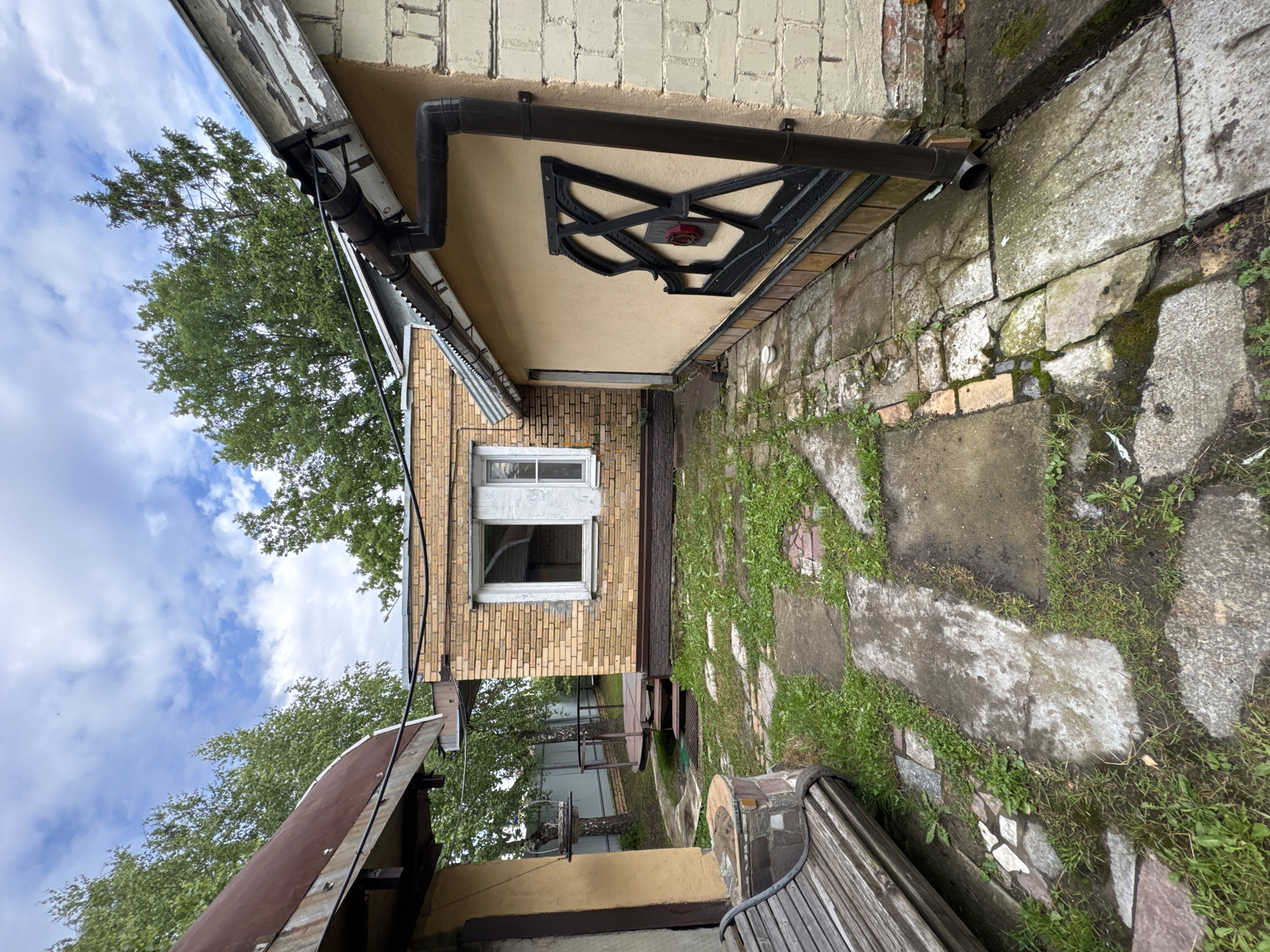 The image shows a slightly overgrown backyard in Naro-Fominsk, Russia. A wooden bench is visible in the foreground, next to a small, stone-clad structure. A pathway made of concrete slabs and some broken tiles leads towards a yellow-colored building with a corrugated metal roof. To the left, a brick building with a light brown facade and a sloping, rusty metal roof is partially visible. Another brick building with yellow bricks and a white window is situated behind the pathway. A dark brown downspout runs along the side of the yellow building. The area is surrounded by trees and a fence in the background. The sky is cloudy with patches of blue.