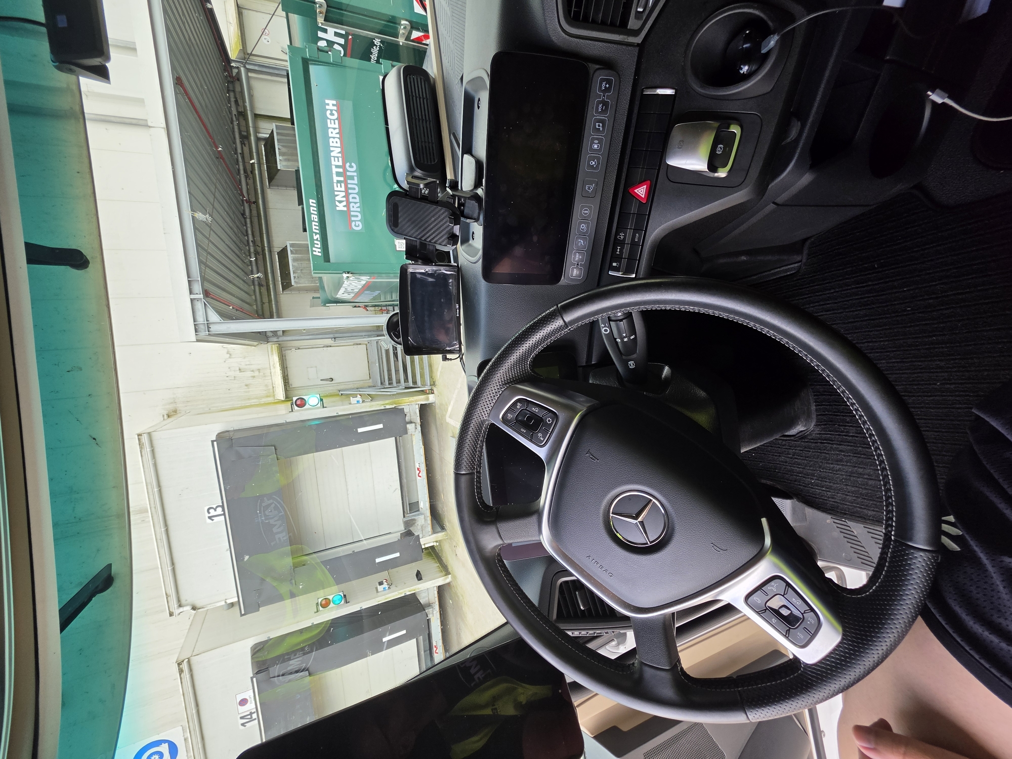 This media file is a photograph taken from inside the cab of a truck, looking out through the windshield and side windows. The interior of the truck is visible, featuring a steering wheel with a Mercedes-Benz emblem, dashboard controls, a digital display screen, and a smartphone mount. A charging cable is plugged into the dashboard.
Outside the truck, the scene appears to be an industrial area or loading dock. There are large, white loading bays visible, some marked with numbers "13" and "14". A green truck or waste disposal vehicle with the text "KNETTENBRECH GURDULIC" and "Husmann" is parked next to the truck. Traffic lights, showing green and red signals, are also visible near the loading bays. The overall environment suggests a daytime setting, possibly a logistics hub or a facility where goods are being loaded or unloaded. The image is rotated 90 degrees counter-clockwise.
