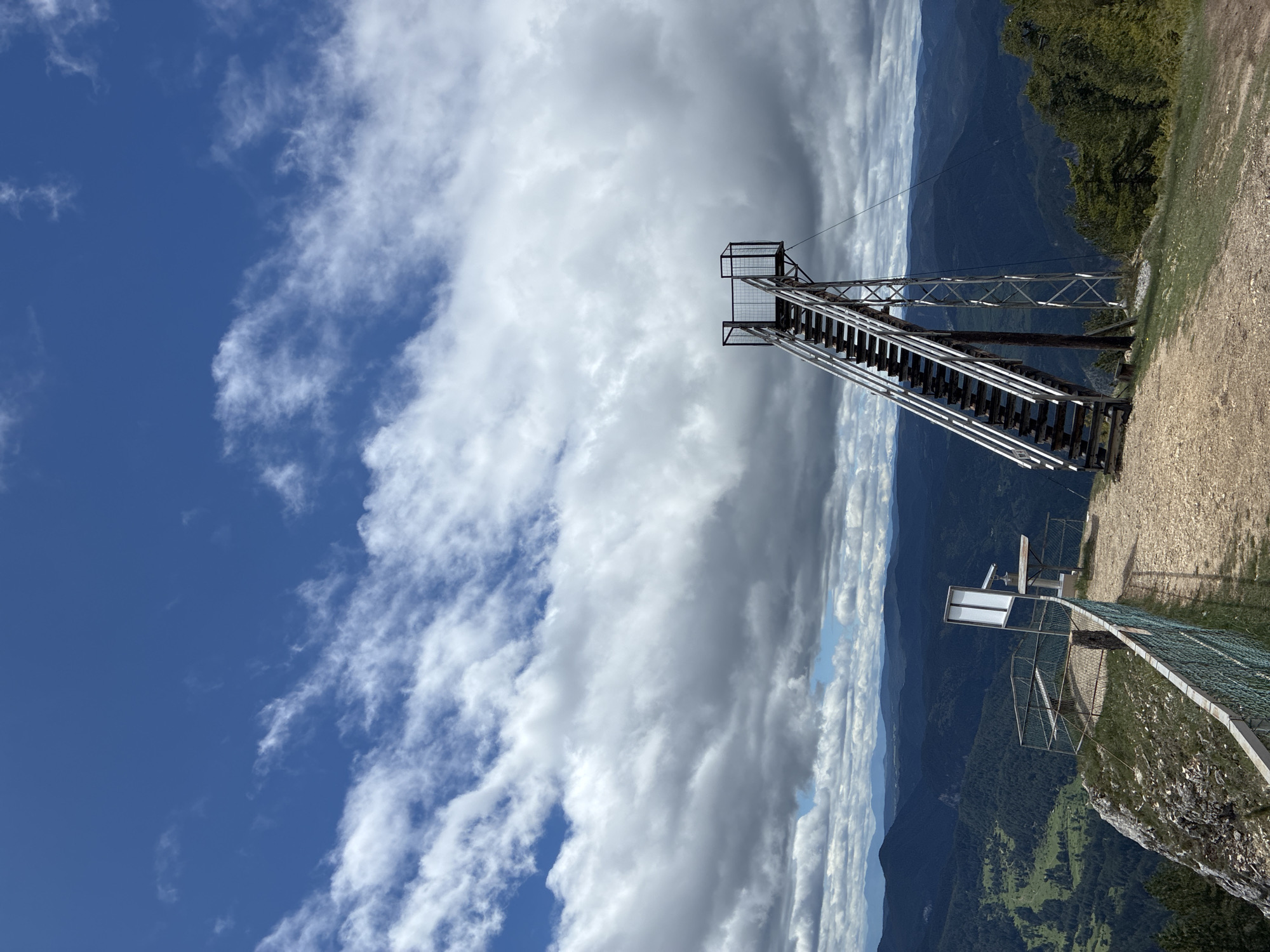 The image depicts a tall metal structure resembling an observation tower on the edge of a cliff or mountain. The tower consists of a staircase leading to a platform at the top. In the background, there's a landscape of mountains and sky with clouds. A portion of the landscape is on the right side of the image, consisting of trees and dirt. A sign is partially visible on the right side of the image. The location is Giovren, Bulgaria.