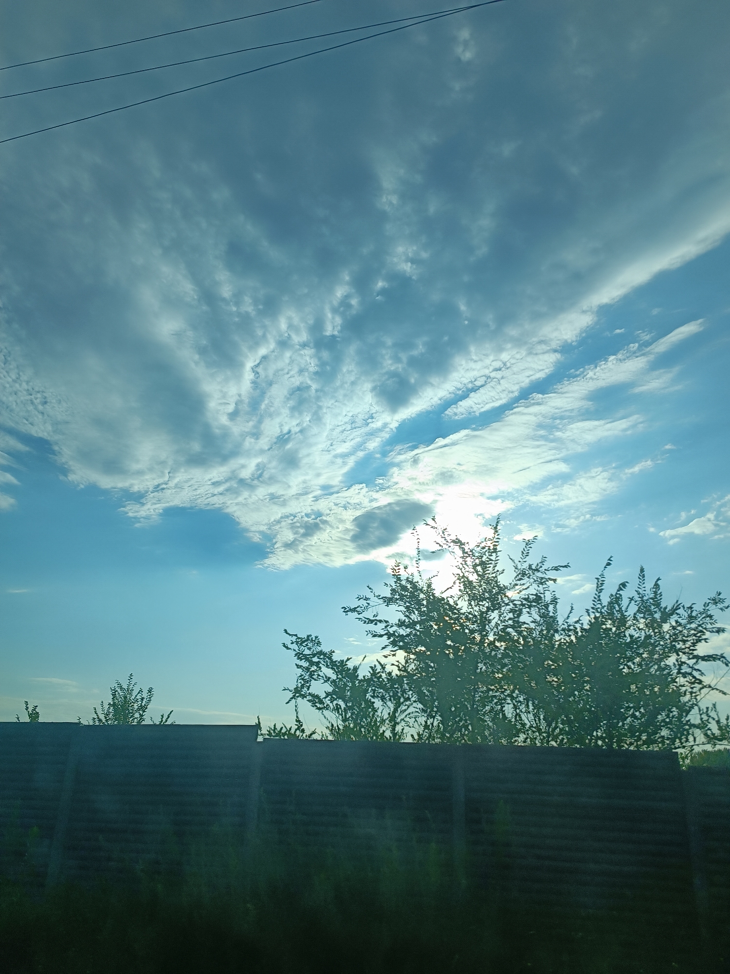 The image shows a dark, horizontally slatted fence in the foreground, with dark green foliage and trees peeking over the top. The sky above is a bright, pale blue, with wispy white clouds scattered across it. Sunlight is visible, partially obscured by some of the clouds, creating a soft glow. Three thin, dark power lines stretch diagonally across the upper portion of the sky. The overall impression is of an outdoor scene, possibly in a residential or semi-rural area, during daytime. The fence appears to be made of wood or a similar material and is a prominent feature in the lower half of the image. The trees are leafy and dense, with some branches extending into the sky.