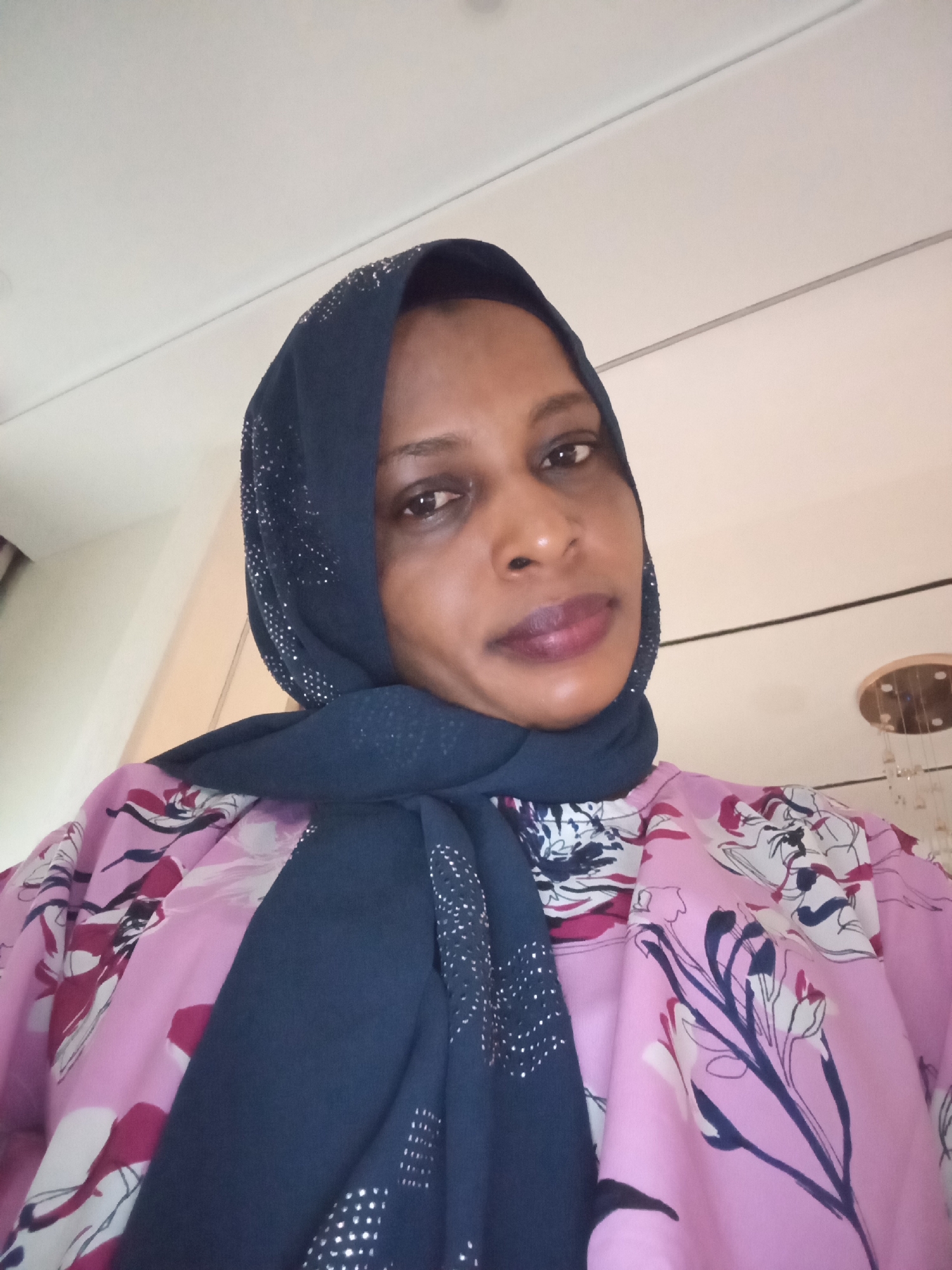 A person wearing a dark blue hijab adorned with small, possibly reflective, embellishments and a light pink shirt with a floral pattern is looking at the camera. The background is simple, with a white wall and a partial view of a ceiling fixture. The location is Unguwar Rimi, Nigeria.