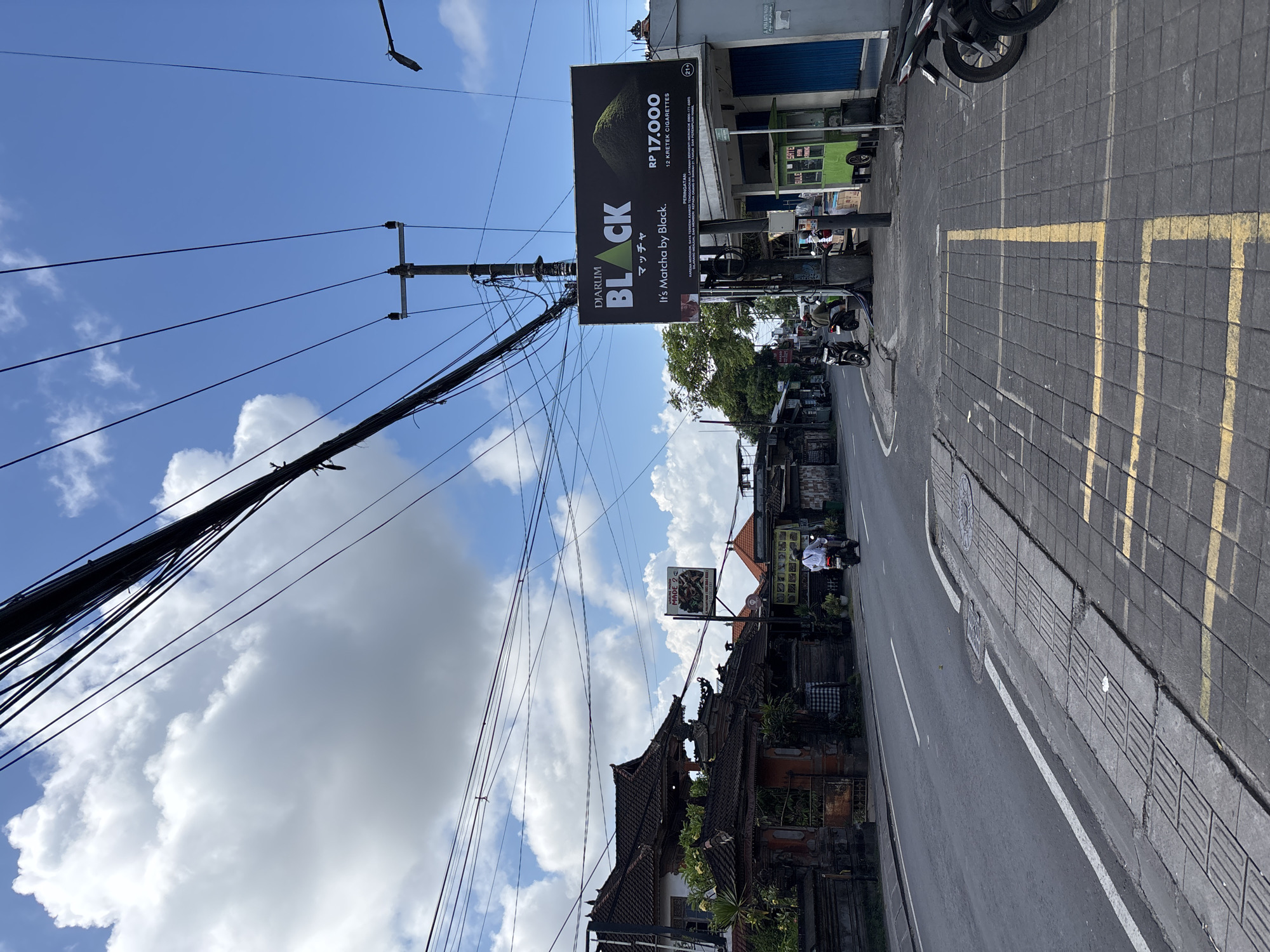 This image depicts a bustling street scene in Kuta Selatan, Indonesia, captured on a bright, sunny day with a clear blue sky dotted with white clouds. The perspective is tilted, showcasing a street with an asphalt road and a grey paved sidewalk featuring yellow rectangular markings.
A prominent utility pole, heavily tangled with numerous electrical wires, stands in the mid-ground. Attached to this pole is a large black billboard advertising "DJARUM BLACK" cigarettes. The text on the billboard reads "マッチャ" (Matcha), "It's Matcha by Black," and shows a price of "RP 17.000" for "12 KRETEK CIGARETTES." It also includes a health warning: "PERINGATAN: LAYANAN BERHENTI MEROKOK 800-177-6500" (Warning: Quit smoking service...) and an age restriction "(21+)."
Along the street, various buildings are visible. On the right, there are shopfronts with blue and green shutters. Further down the road, traditional Balinese architecture with dark, ornate tiled roofs and carved details can be seen. Several motorcycles are present, some parked on the side of the road, and others being ridden by individuals. Another smaller sign on a building reads "MADE 20," while other partial texts like "SATE," "ULE," and "ARJUNA" are also visible. The image conveys the lively atmosphere of a typical street in an Indonesian town.