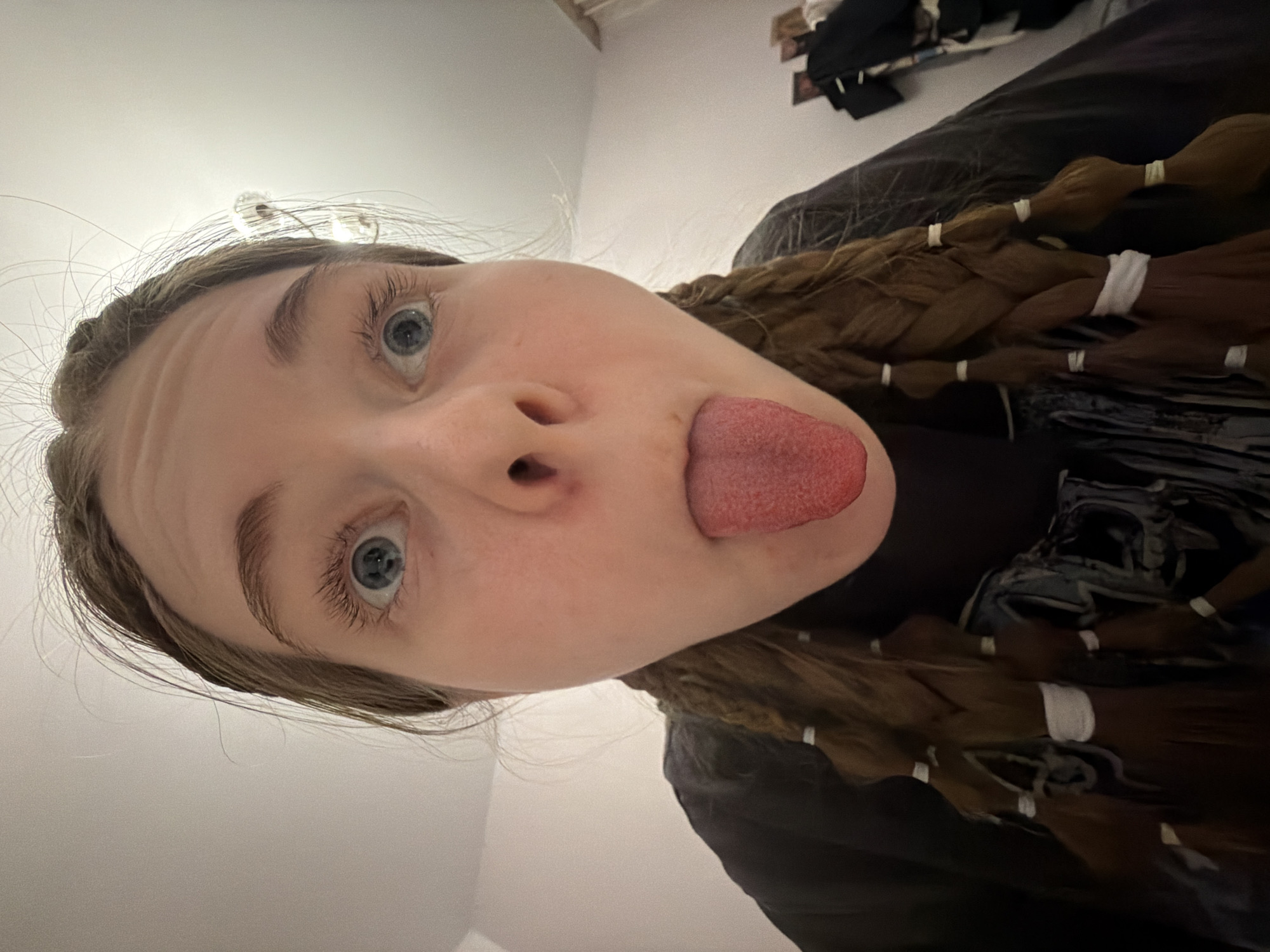 A young person, likely female, with fair skin, wide blue eyes, and light brown hair, is captured in a close-up selfie taken from a low, upward-looking angle. The subject's face is prominent in the frame, with her tongue sticking out in a playful or silly expression. Her eyes are looking directly upwards towards the camera.
Her light brown hair is styled in multiple thick braids, which are secured at intervals with small white elastic ties or bands. These braids are visible along the right side of the image, resting against a dark, possibly fabric-covered, surface. She appears to be wearing a dark-colored top, partially visible around her neck and shoulder.
The background is a plain, light-colored wall, possibly white or off-white. In the upper right corner, there is a glimpse of what appears to be a dark-colored bed or sofa with some items on it or a wall shelf above it, suggesting an indoor setting. The overall mood conveyed by the expression is lighthearted and humorous. There is no visible text in the image. The image is set in Perm, Russia.
