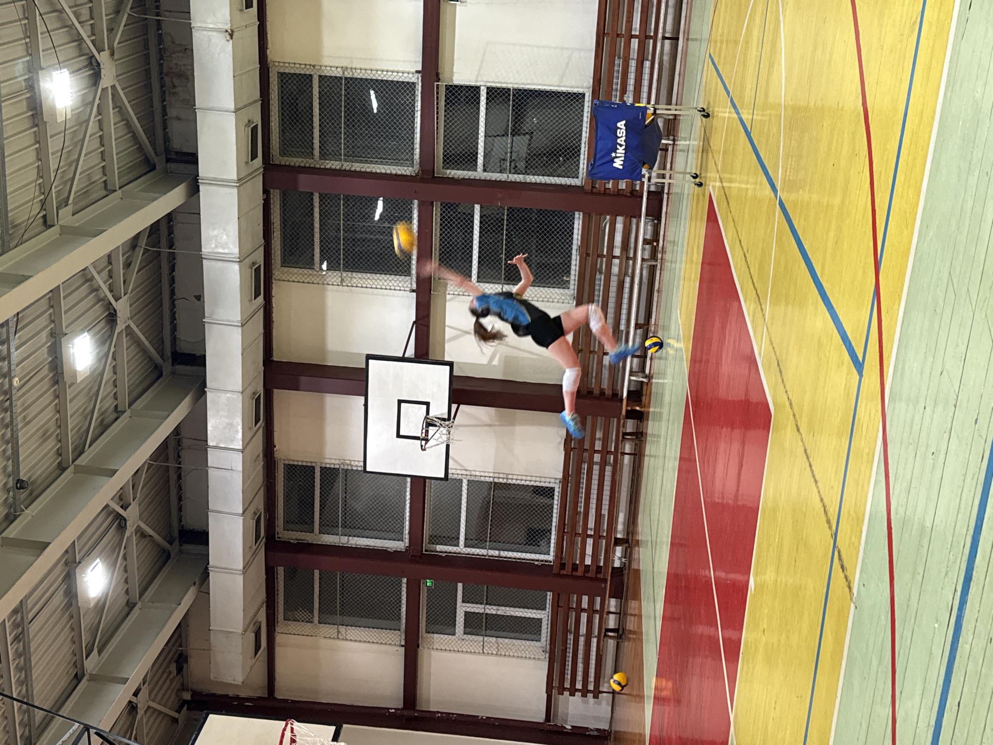 The image shows an indoor gymnasium. A woman is jumping mid-air to strike a volleyball. There's a basketball hoop affixed to the wall. The gymnasium has a wooden floor with red, blue, and yellow lines. Metal-framed windows with wire mesh are visible on the walls. A Mikasa volleyball net stand is present near the edge of the playing area. The ceiling has metal structures and lighting fixtures. The exact location within Sofia, Bulgaria, cannot be determined from the image.