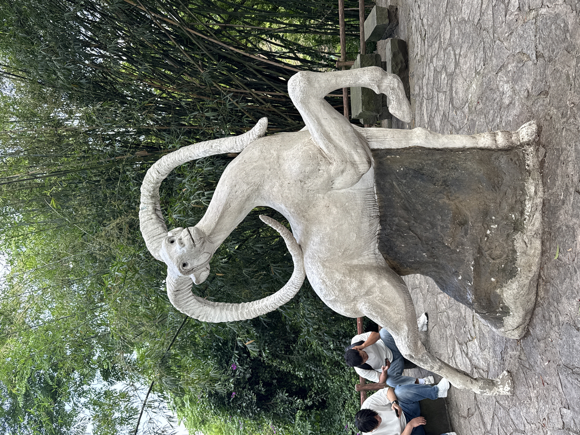 The image shows a concrete sculpture of a horned animal in a park. Two people are sitting on a bench nearby, with their heads facing away from the camera. They are wearing white shirts and blue jeans and appear to be occupied with their phones. The sculpture is positioned on a stone platform or base. The background consists of dense green foliage, including bamboo trees. The setting is possibly a park or garden setting, based on the vegetation and stone pathway. Based on the question prompt, the location is within Jiulongpo District, China.
