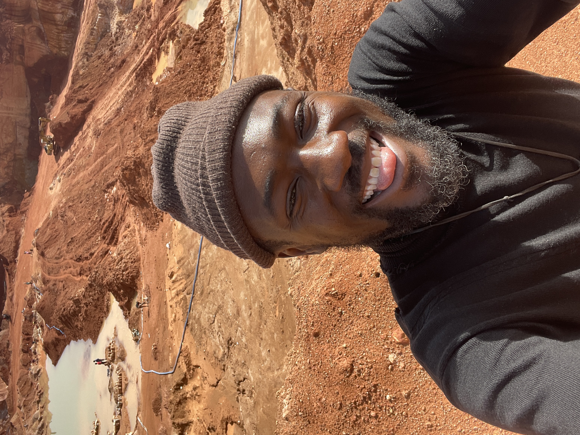 A smiling Black man wearing a brown beanie and black long-sleeved shirt is photographed at an angle above what appears to be a mining site in Jos, Nigeria. The landscape features red soil, pools of water, and heavy machinery. Some figures can be seen working near the pools of water. A long blue pipe stretches across the landscape. The man's face is illuminated by sunlight. His mouth is open, revealing his teeth. His beard is short and graying.