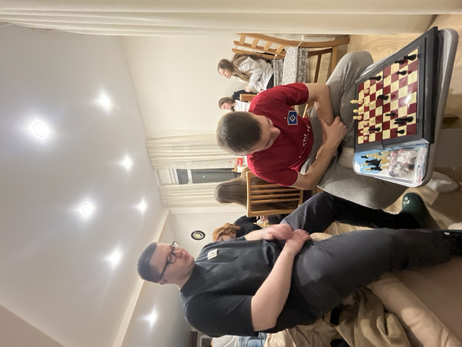 The image displays an indoor scene located in Заозерное Снт, Russia, depicting a social gathering. In the foreground, two males are prominent. The male on the left, wearing spectacles, a black short-sleeved t-shirt, and dark trousers, is seated on the edge of a light-colored sofa, looking downwards with his hands clasped. He wears a dark green slip-on shoe on his visible right foot. The male on the right, clad in a red short-sleeved t-shirt featuring a white Hamburg SV logo and light grey sweatpants, is seated and intently focused on a chess board placed on a small table before him. The chess board has red and off-white squares with both black and white pieces actively set for play; additional chess pieces are contained in a clear plastic bag next to the board.
In the mid-ground and background, approximately five other individuals are visible, primarily seated at a wooden dining table with chairs. A woman with reddish-brown hair is partially visible to the left of the table. A red Coca-Cola bottle rests on the table. The room features light-colored walls, a wooden floor, and a white ceiling equipped with multiple recessed downlights. A large window with white curtains is visible in the background, showing a dark outdoor environment. A round, dark object is mounted on the far wall.