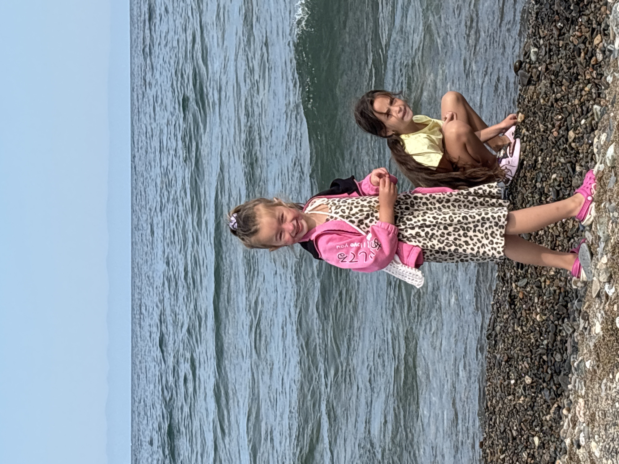 Two young girls stand on a rocky shore near the water's edge. The girl in the foreground, on the left, is wearing a pink hooded jacket over a leopard-print dress and pink Crocs. She has her hair pulled up in a ponytail and is smiling at the camera. The girl in the background, on the right, is crouching down and looking towards the water. She is wearing a yellow t-shirt and pink Crocs. The water is a dark blue with small waves, and in the distance, a hazy line of mountains can be seen against a light blue sky. The rocky shore is covered in dark gray and white pebbles. The location is identified as Rechka Mishikha, Russia.
