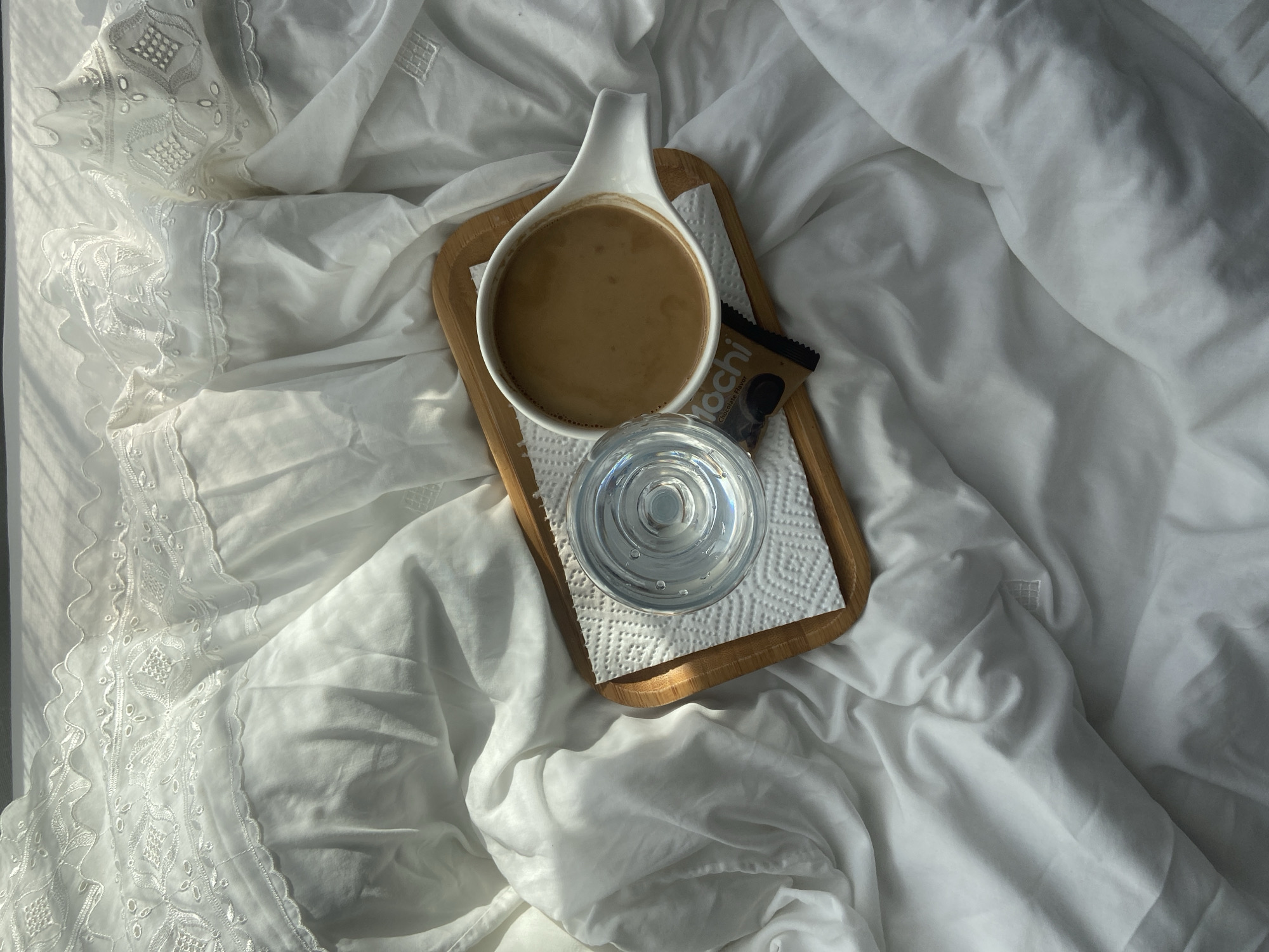 The image presents a still life composition on a bed covered with white linens, possibly located in a hotel room or a private residence. A light-colored wooden tray is positioned on the bed. On the tray, there is a white cup filled with a coffee-colored liquid, next to a sealed chocolate mochi, and a glass of water on a white napkin. The linens are creased and wrinkled, suggesting a relaxed or recently occupied setting. The lighting is soft, with some areas in brighter light and others in shadow, which adds depth and texture to the scene. The setting is serene and evokes a sense of comfort.