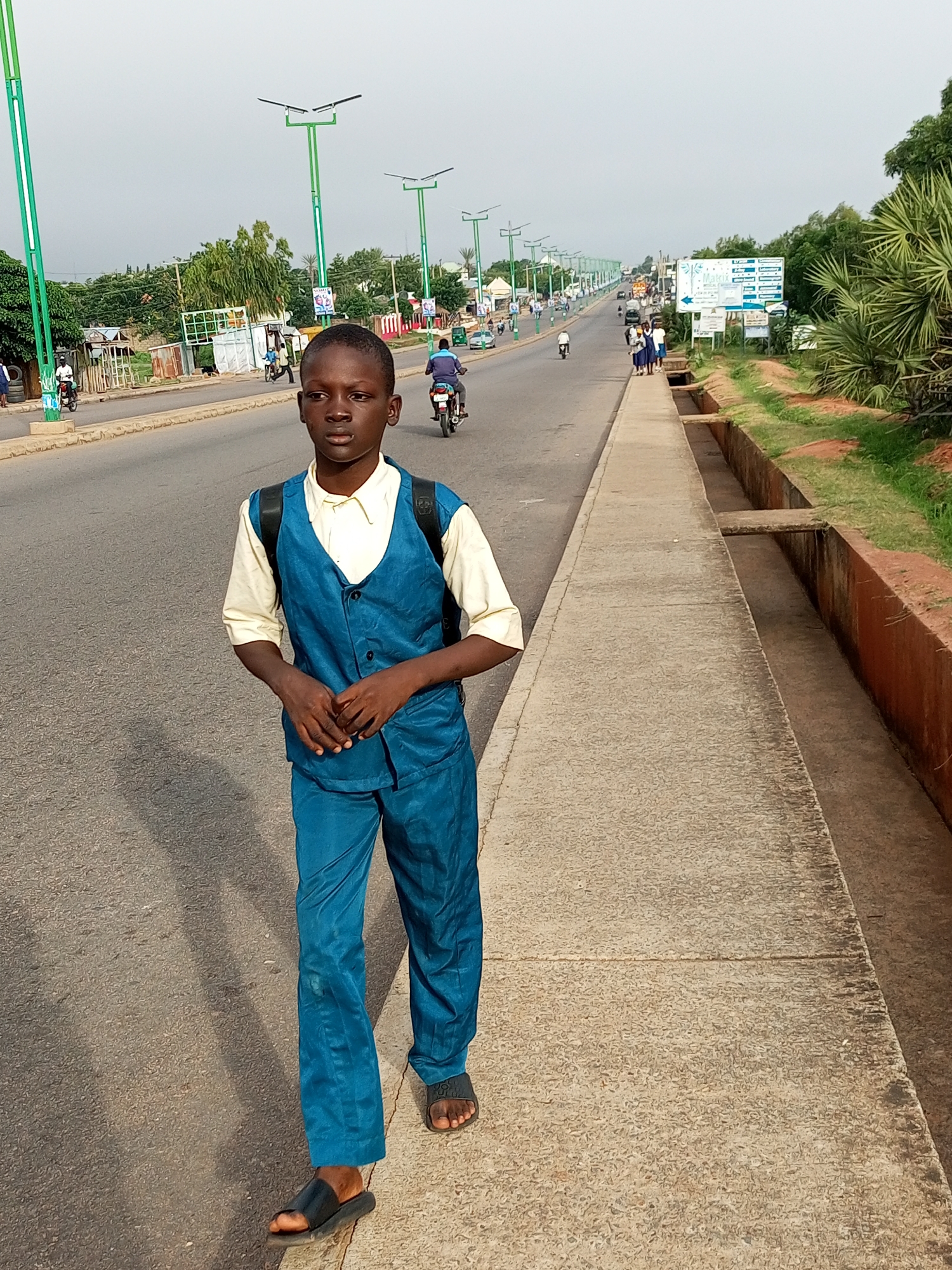 The image captures a young boy walking on a sidewalk next to a road in Garko, Nigeria. He is wearing a blue vest and matching pants over a light-colored shirt, with a black backpack on his shoulders. The road is paved and shows traffic, including motorcycles and cars, with streetlights lining the side. The sidewalk is made of concrete, and a drainage ditch runs alongside it. In the background, buildings and vegetation are visible, suggesting an urban or semi-urban environment. The sky is overcast.