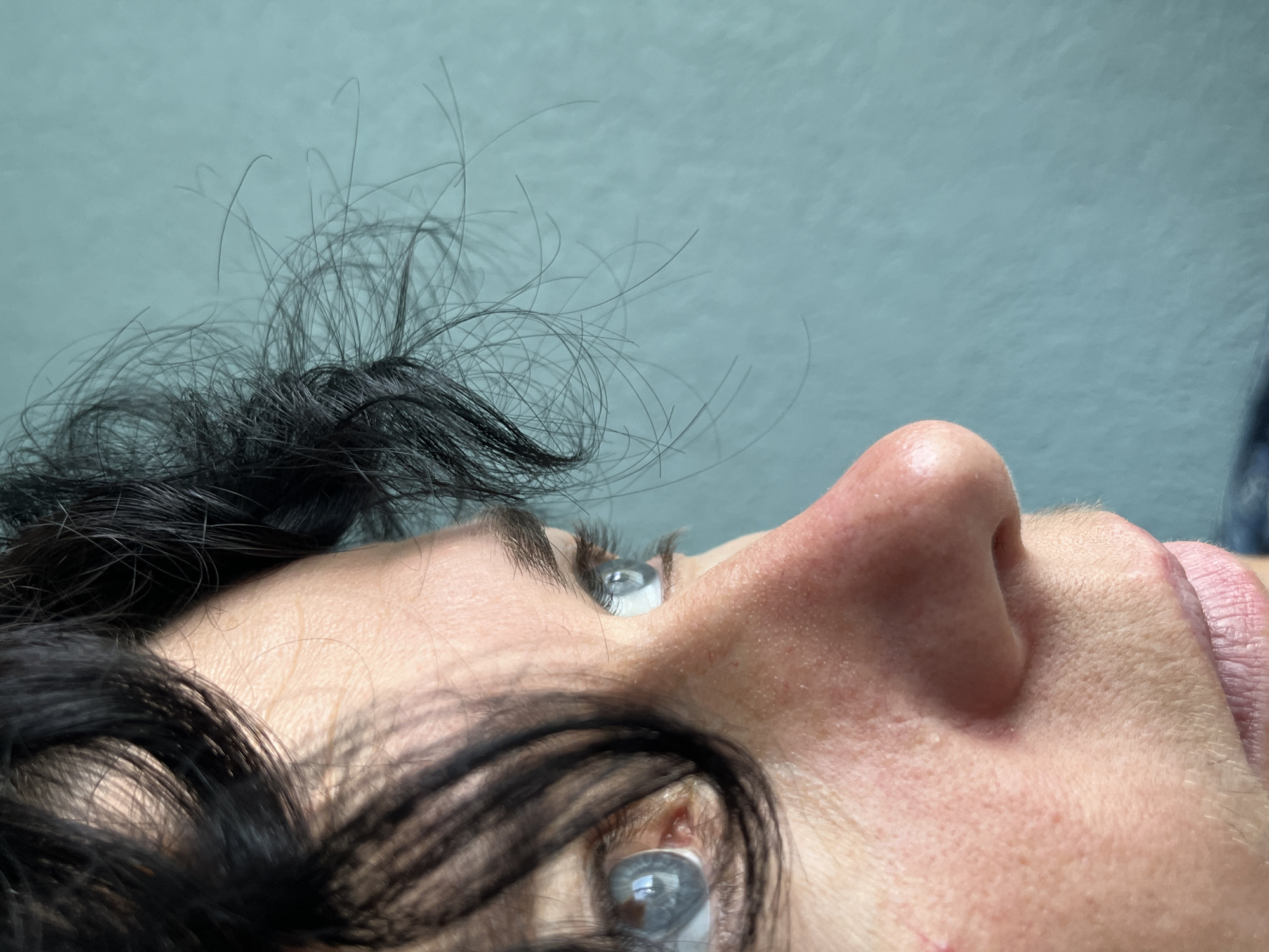 The image is a close-up profile shot of a person's face, focusing on their left eye, nose, and mouth. The person has dark, curly hair that partially obscures their face. Their eyes are a striking blue-gray color, with visible eyelashes. The skin appears fair with some texture and slight redness on the cheek. The background is a solid, muted teal color. There is no indication of a specific location within the city of ღრმაღელის თემი, Georgia, as the image solely captures the subject's face and a plain backdrop.