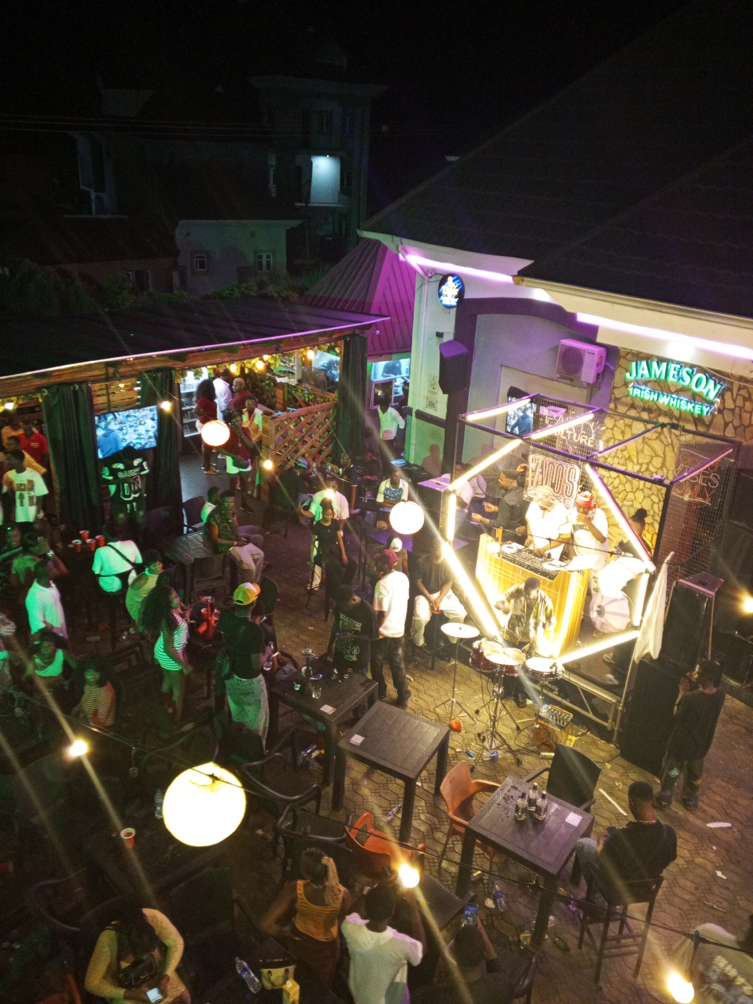 The image shows an elevated view of an outdoor bar or entertainment venue, likely located in Lokoja, Nigeria. Several people are gathered, suggesting an active social scene. There is a live band performing on a stage that has a drum kit. The band and DJ equipment are illuminated by neon lights and a sign on the back of the stage reads "Vados" - an apparent advertisement for the bar. Some patrons are sitting at tables, while others are standing and socializing. There are mounted televisions and string lights, indicating an effort to create a festive atmosphere. A Jameson Irish Whiskey sign is attached to one of the buildings. A building in the background suggests an urban setting.