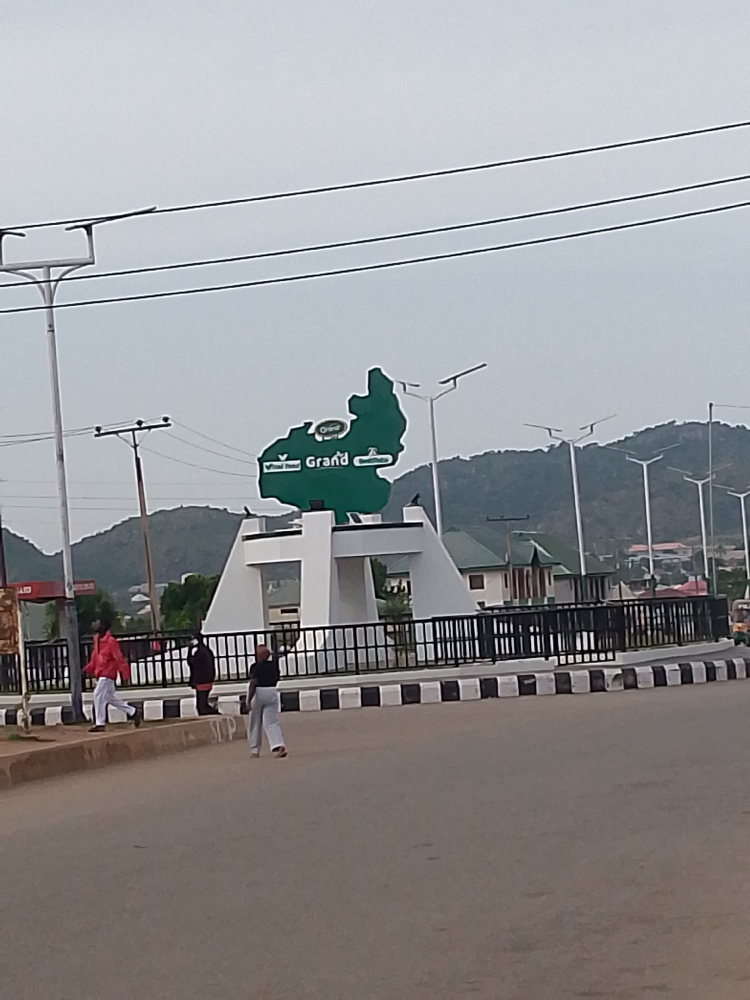 The image shows a green landmark in the shape of a map with the word "Grand" written on it, located in Kuru Dungus, Nigeria. The landmark is situated on a white, multi-tiered structure, which is surrounded by a black metal fence. In front of the fence, there is a road with black and white striped borders. Three individuals are visible walking on the sidewalk and road. One person in a red jacket is walking towards the left, a person in a black outfit is walking in the middle, and a person in a white outfit is walking towards the right. In the background, there are several streetlights, buildings with green roofs, and lush green hills under a cloudy sky. Overhead power lines are also visible.