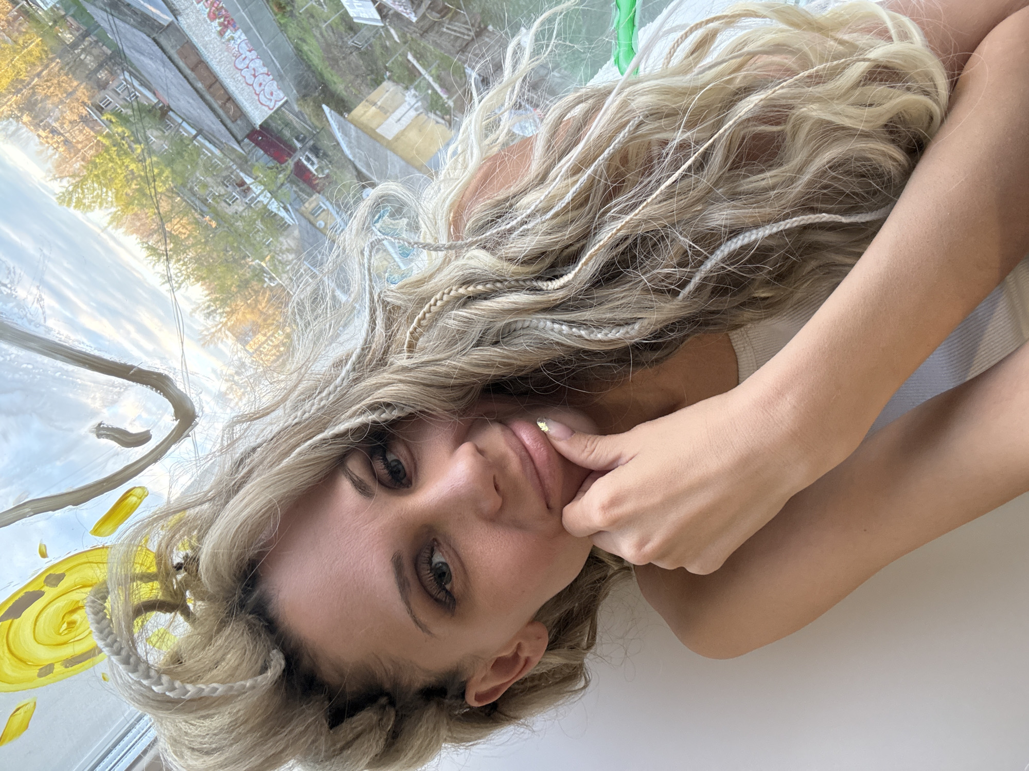 A young woman is the central subject of this image, captured in Rusanovo, Russia. She is shown lying down, with her head tilted, looking directly towards the viewer. Her long, wavy blonde hair, interspersed with several fine, light-colored braids, cascades around her. She wears a simple, light-colored (possibly white) ribbed top. Her right hand is brought up to her face, with a fingertip gently touching her chin or lower lip, revealing a small gold glitter detail on her fingernail. Her expression is calm and contemplative, with a faint, subtle smile.
The setting places her indoors, seemingly next to or lying on a surface beside a window. The window pane itself is creatively adorned with artistic, abstract designs painted in yellow and white, featuring a prominent spiral shape and various brushstrokes. Through this decorated window, an urban or residential outdoor scene is visible. This background depicts several multi-story buildings, some exhibiting graffiti on their walls, including partially visible Russian letters such as "СКУ." Lush green trees and others with autumnal yellow foliage are scattered throughout the landscape. The sky is partially visible, indicating daytime, with soft light suggesting a late afternoon.