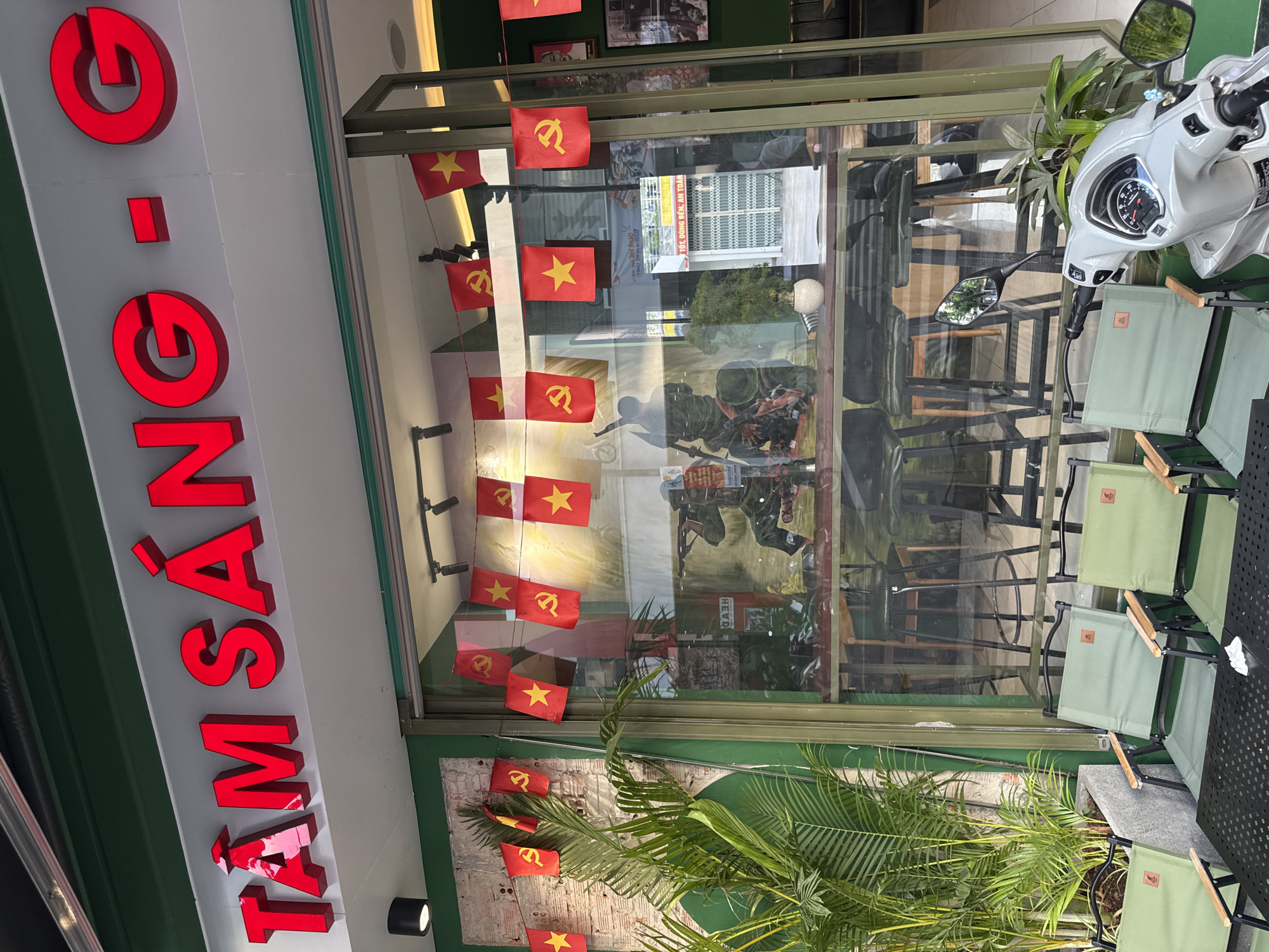The image depicts the storefront of a business in Nha Trang, Vietnam, identified by the text "TÂM SÁNG - G" in large red neon letters above the entrance. The storefront features a large glass window revealing the interior, which appears to be a cafe or restaurant with bar stools and tables. Small red flags with yellow stars and hammers and sickles, representative of the Vietnamese flag and communist symbolism, are strung across the inside of the window. In front of the storefront, there are several light green camping chairs and a black table, suggesting an outdoor seating area. A silver scooter is parked to the right of the entrance, partially obscuring the view of the chairs and table. Lush green tropical plants are visible to the left of the entrance. The exterior of the building is painted green. The overall impression is a commercial establishment with a clear nationalistic and possibly communist theme.