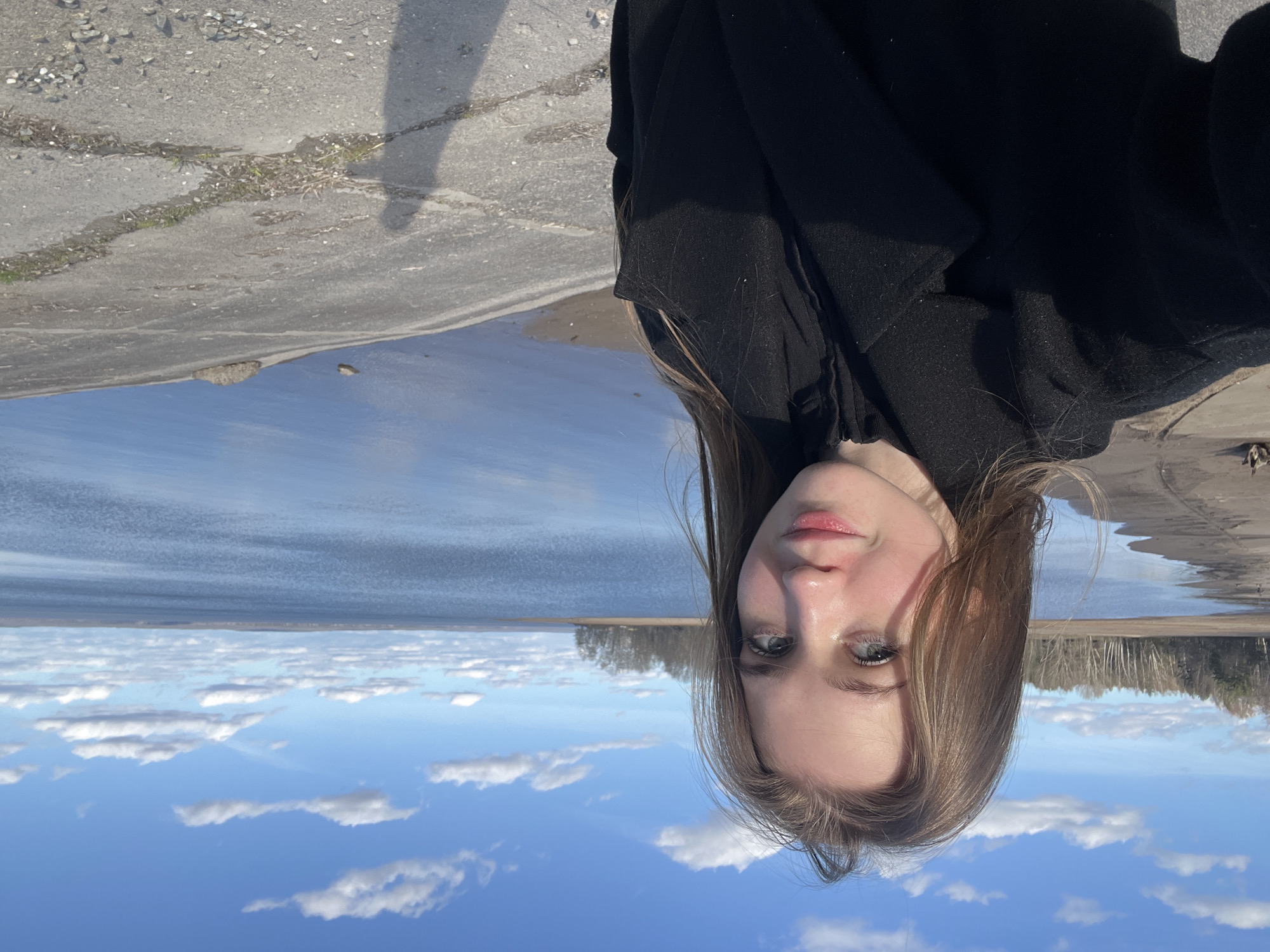 A portrait-style photograph, rotated 180 degrees, shows a young woman in a black jacket, against a backdrop of a cityscape.
The upper portion of the image features a blue sky with scattered white clouds. Below this is a horizon line, indicating a body of water or the sky reflected in the water. The cityscape is faintly visible along the horizon, appearing as a line of trees or buildings.
The woman is positioned in the foreground, with her face clearly visible. Her hair is light brown, and her skin is fair. She looks directly at the camera. The black jacket she is wearing takes up a portion of the top of the frame, partially obscuring the environment behind her.
A concrete structure is visible in the top left corner, possibly a ledge or part of a building. A dark shadow is also cast on this concrete area. The location is Noviy Urengoy, Russia, which is implied by identifying the objects and subjects.
