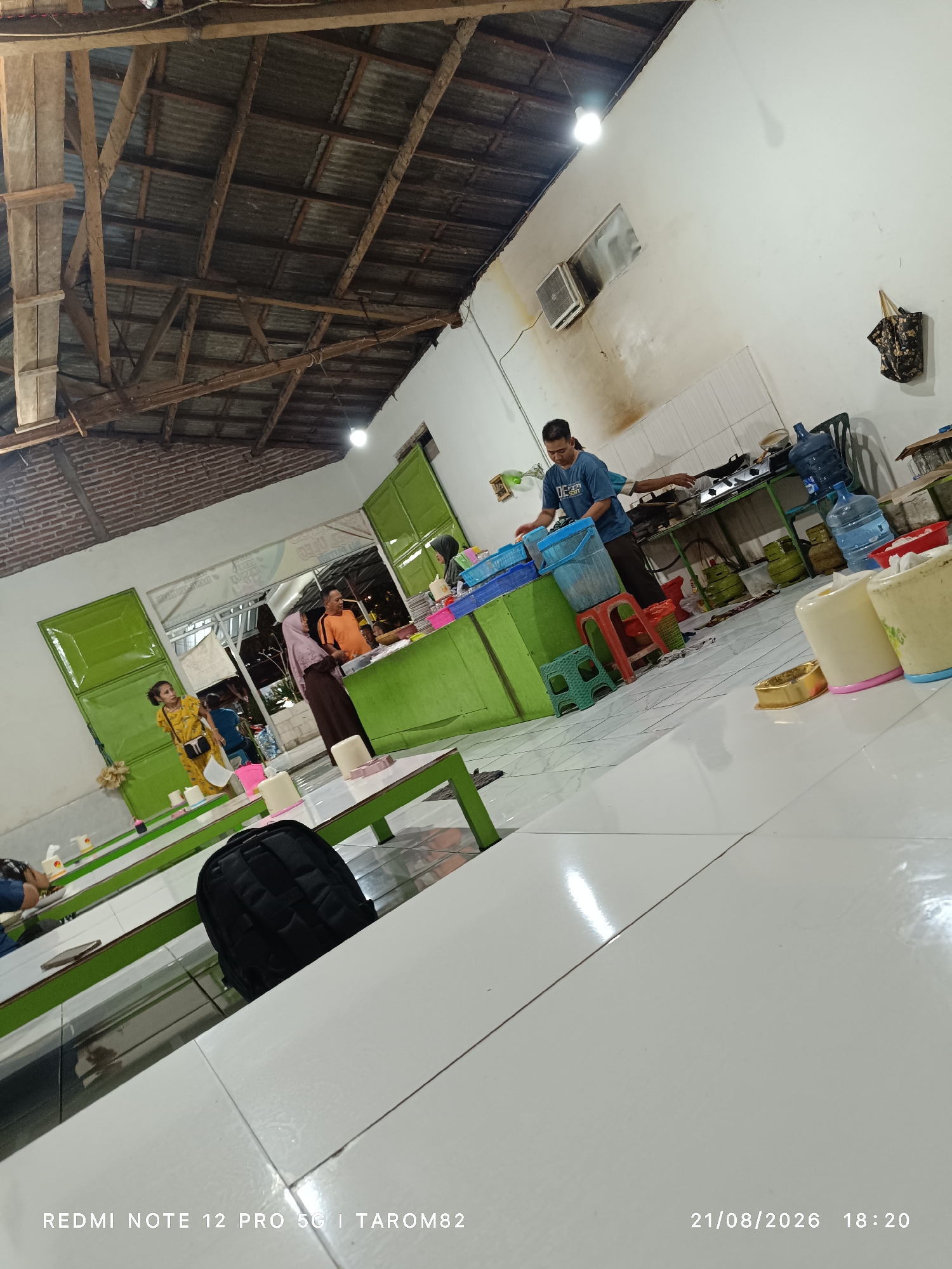The image depicts an indoor scene, likely a simple eatery, in Sidodadi, Indonesia. The setting features a rustic wooden ceiling with exposed beams and corrugated metal roofing. The walls are white, with some areas showing signs of staining and discoloration. The floor is tiled in white.
In the background, a man in a blue t-shirt is standing near a stove, seemingly engaged in food preparation. Various cooking utensils, gas cylinders, and large water bottles are visible on a counter behind him. Several people are milling about in the open doorway and within the establishment.
In the foreground and mid-ground, green tables with white tops are set up, some with cups and other small items on them. A black backpack rests on one of the tables. A woman in a yellow dress and a woman in a hijab are standing near the tables, and another woman in a dark brown hijab and an orange shirt stand near a green counter. The overall impression is of a casual, community-oriented dining space. The timestamp in the lower right corner indicates the image was taken on August 21, 2026, at 18:20.