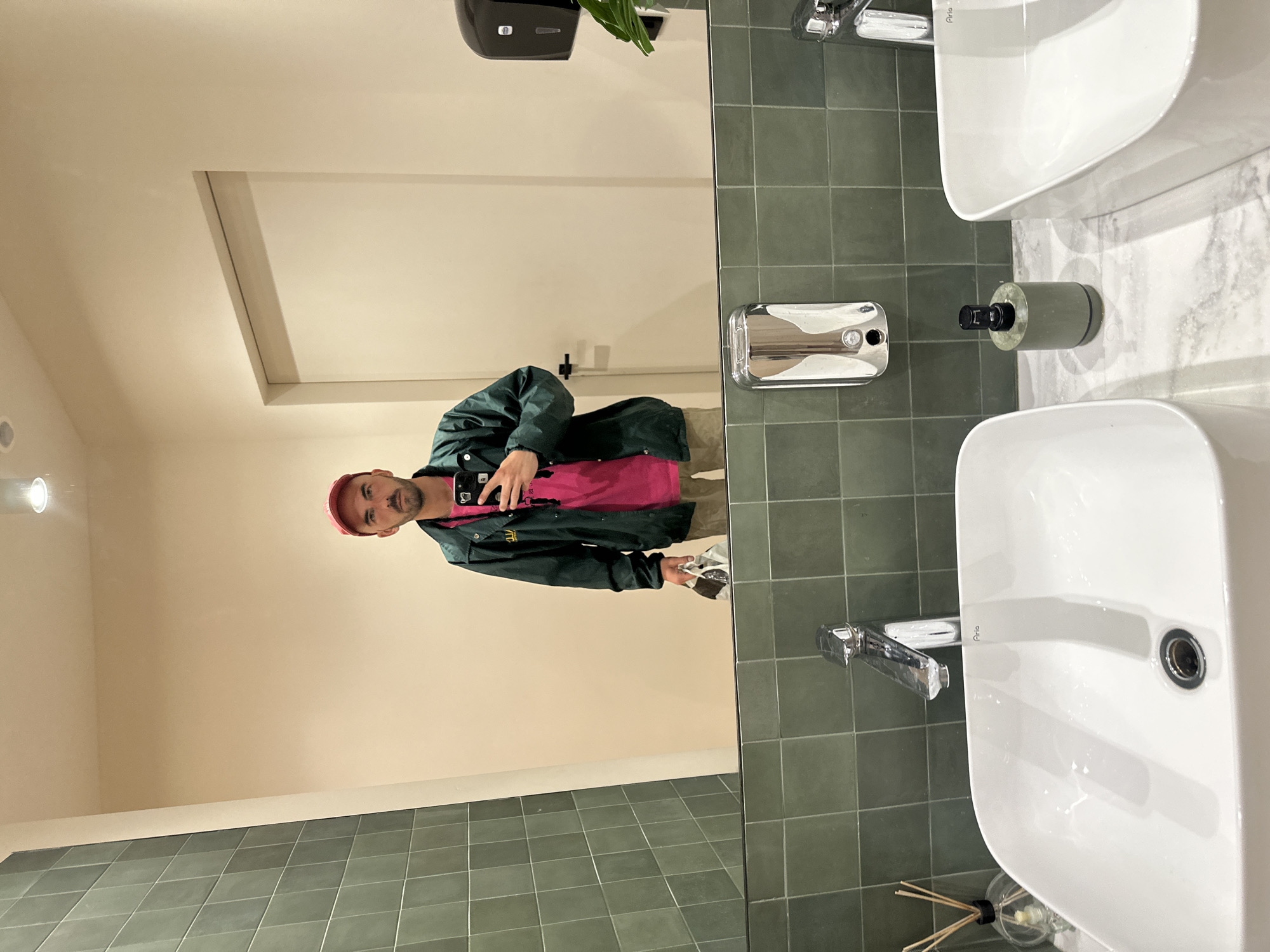 The image shows a reflection in a mirror in a bathroom. The reflection captures a person with fair skin, a red cap, and a dark green jacket over a pink shirt, holding a phone in their left hand and some papers or a napkin in their right hand. The bathroom has green square tiles on the wall and floor, and two white sinks are visible, with chrome faucets and soap dispensers. The wall behind the person in the reflection is a light beige color. The photo appears to be taken in a public restroom, possibly in Budva, Montenegro, based on the style of the bathroom fixtures.