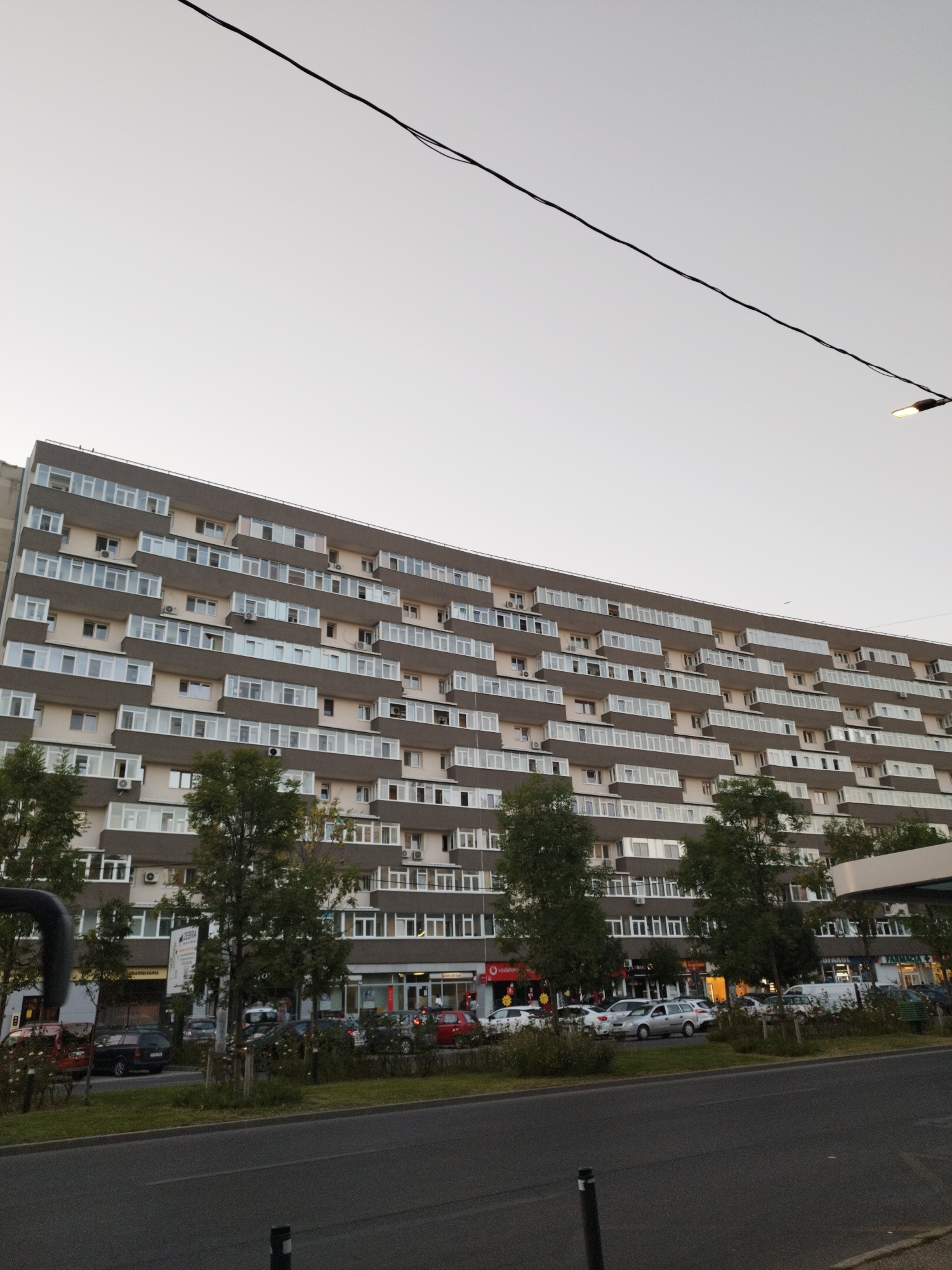 The image shows a long, multi-story residential building in Bucharest, Romania, possibly a block of flats. The building has a modern design with many balconies, each enclosed with glass windows, creating a uniform facade. The upper floors feature rows of windows and air conditioning units. In front of the building, at street level, there are several businesses with storefronts and illuminated signs, including what appears to be a "Vodafone" store and a "Farmacia" (pharmacy). Trees line the sidewalk in front of the shops. Several cars are parked along the street and in front of the businesses. The sky is overcast and light grey. A thin black cable stretches across the top of the frame, and a street lamp with its light on is visible in the upper right corner. The scene depicts an urban environment on a slightly overcast day.