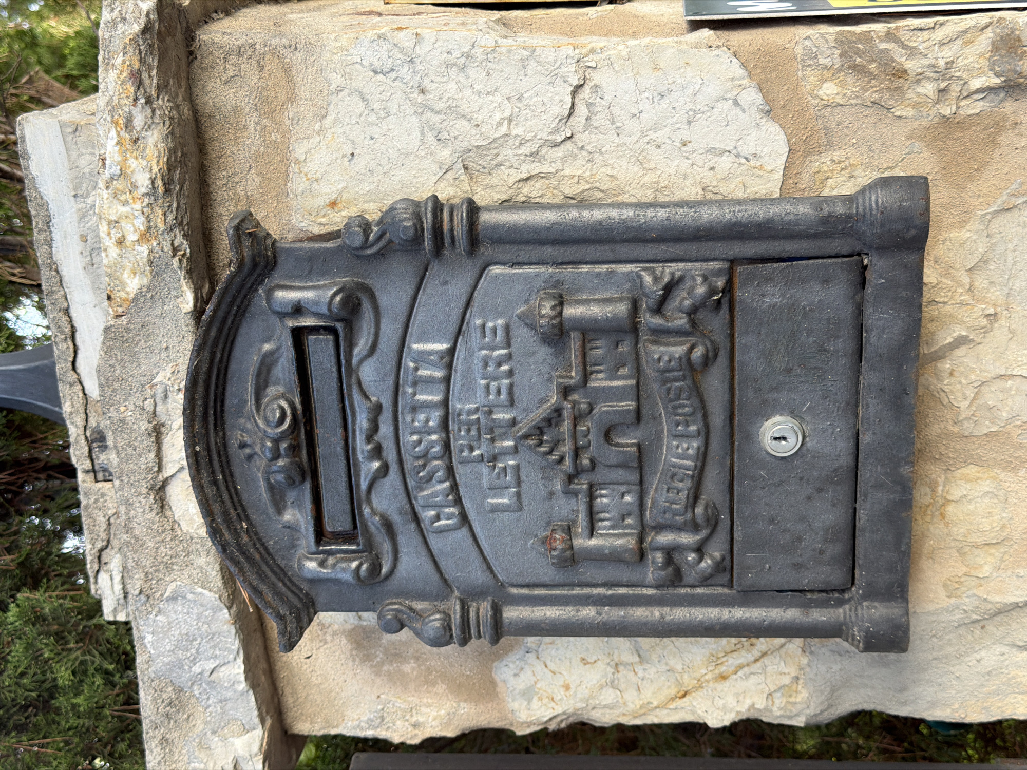 A black cast iron mailbox is mounted on a stone pillar. The mailbox has decorative carvings, including a castle and the words "CASSETTA PER LETTERE" and "REGIE POSTE". There is a small keyhole below the decorative carvings. The pillar is made of rough-hewn stone and is surrounded by greenery. The mailbox is in the city of Palmanova, Spain.
