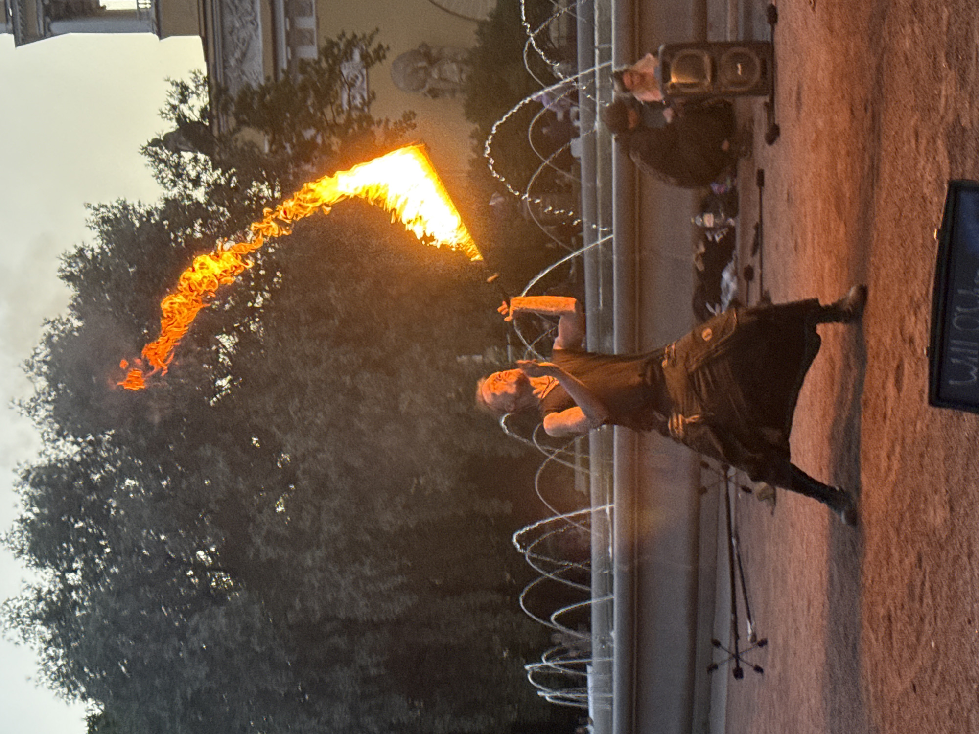 A male fire performer, dressed in dark clothing and a flowing skirt, is captured in mid-performance, spinning a prop that is emitting a long trail of fire, against the backdrop of a fountain in St. Petersburg, Russia. The setting appears to be an outdoor public space, possibly a park or square, with trees and architectural elements visible in the background. The sky is overcast, suggesting late afternoon or early evening. To the right of the performer, two individuals are seated on the ground near a speaker, observing the performance. Various fire performance props are scattered on the ground around the performer, indicating preparation or a break in the show. The ground is covered with a reddish-brown sandy material. The overall scene depicts a dynamic and captivating street performance in an urban environment.